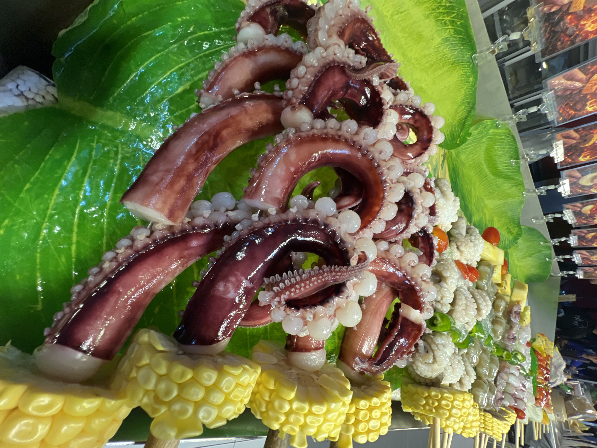 At a street food stall in Karon, Thailand, fresh octopus tentacles are displayed on large, bright green artificial leaves, arranged in a visually appealing manner. The tentacles are a deep reddish-purple, with rows of white suckers clearly visible along their length. Interspersed with the octopus are skewers of corn on the cob, cut into manageable chunks and arranged vertically. In the background, other food items, possibly various seafood or meats, are displayed on skewers, and in the distance, blurred figures of people can be seen, suggesting a busy market environment. The overall scene captures a vibrant and enticing presentation of fresh ingredients ready for grilling or other preparation.
