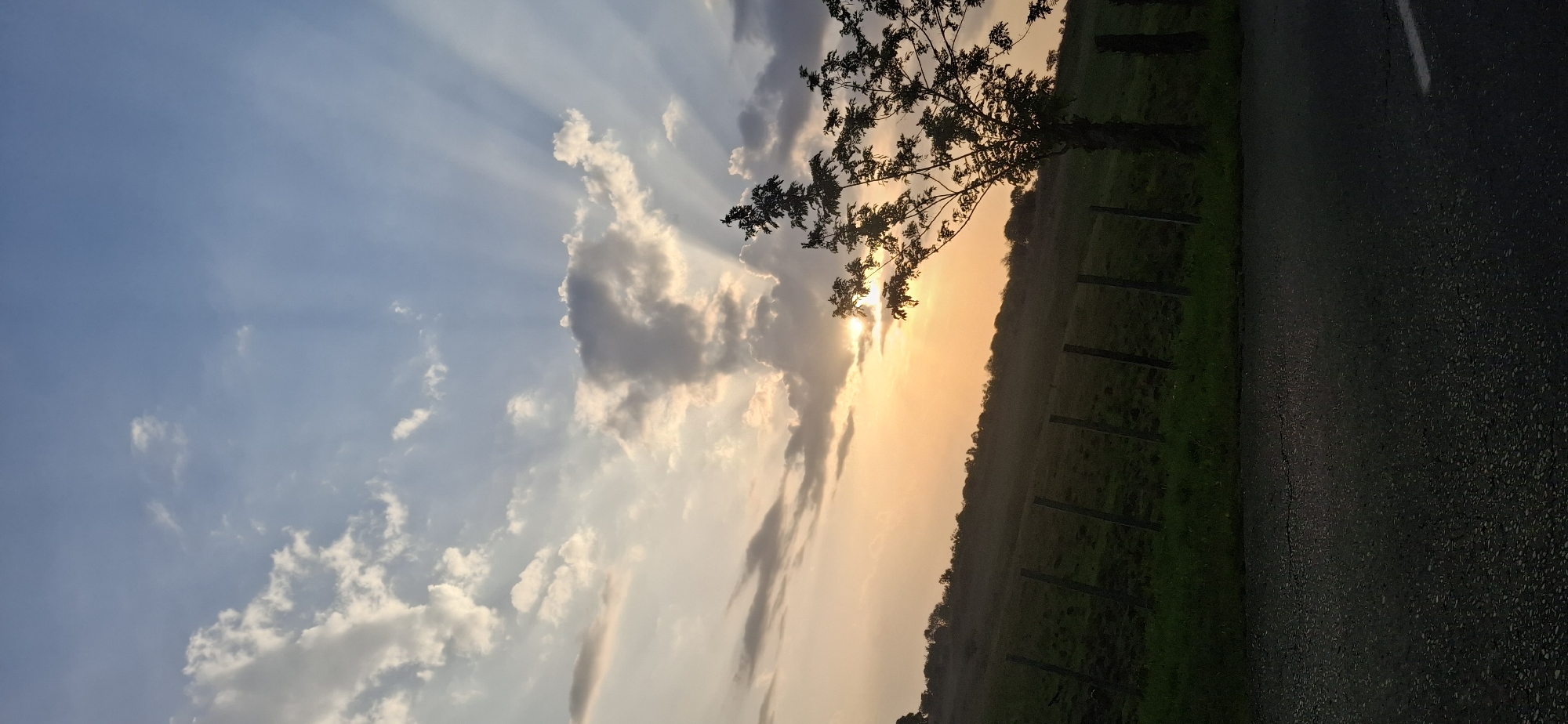 The image shows a sunset scene, likely near Vallecito, Venezuela, though specific details to confirm the exact location are absent. The sun is obscured by clouds and a tree, creating a silhouette effect. Sunbeams are visible through the clouds. To the right of the image is a field with fence posts and a paved surface suggesting a road. The sky transitions from blue to orange and white as the sun sets.