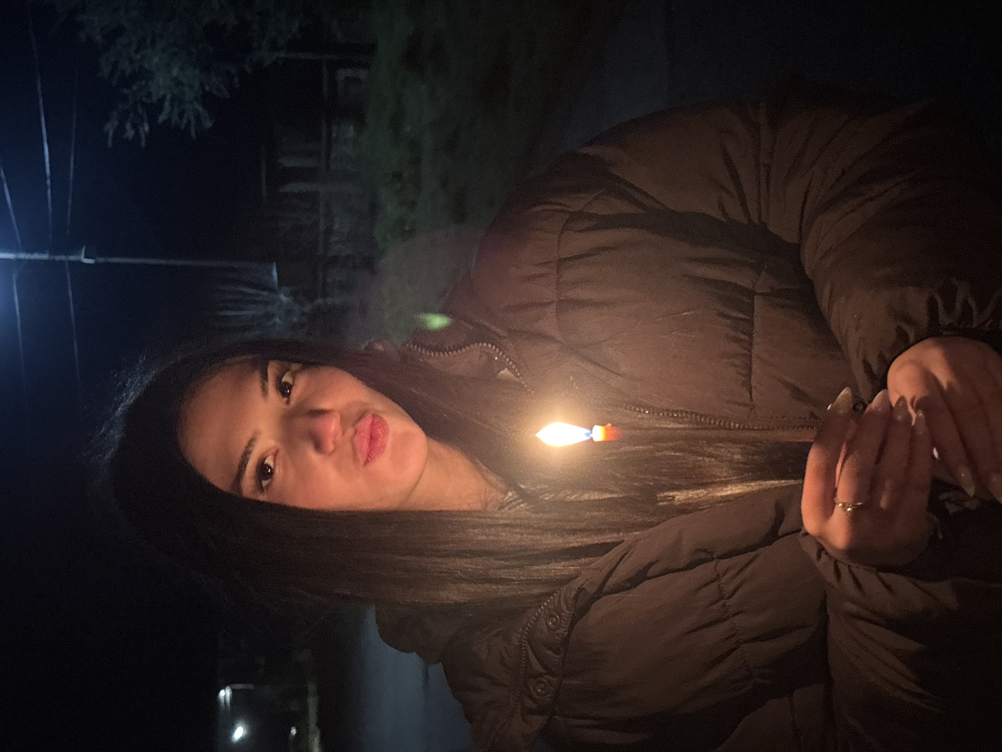 A woman is outdoors in Lehchevo, Bulgaria, at night. She is holding a lit match. She is wearing a dark-colored, puffy jacket and a ring is visible on her right hand. Street lighting is faintly visible in the background behind a dark tree.