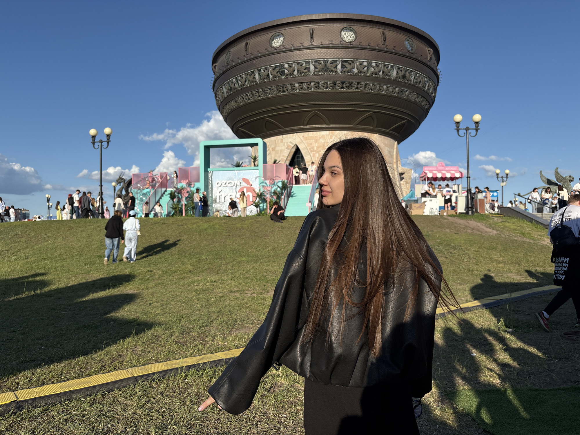 This outdoor daytime image captures a scene in Kazan, Russia, centered around the Kazan Family Center (also known as "The Chasha" or "Cauldron"). The sky is clear blue with scattered white cumulus clouds.
In the immediate foreground, a young woman with long, straight, dark brown hair is depicted with her back partially turned, looking over her right shoulder towards the left. She is wearing a black, loose-fitting, leather-like jacket over a dark garment. Her right arm is extended downwards and slightly forward, with her index finger pointing towards a yellow and black ribbed cable protector or boundary strip laid across the green grass. Long, dark shadows of multiple individuals, including the main subject, are cast onto the grassy slope, suggesting strong, low-angle sunlight.
The mid-ground consists of a grassy hill populated by numerous individuals of varying ages, casually dressed, either standing, walking, or sitting. Several black lampposts with two spherical lights each are interspersed across the scene. Various temporary structures for an outdoor event are visible. These include a prominent light teal rectangular frame with a cascading water feature and partial text "OZON Fresh," along with associated pink and teal platforms, steps, and decorative elements resembling palm trees. A dark, dragon-like statue is positioned near the teal frame. To the right, a smaller, pink and white striped tent-like booth is present, and further right, a large, winged statue or monument stands on an elevated grassy section.
The background is dominated by the distinctive Kazan Family Center, a large, bronze-toned, bowl-shaped building featuring intricate patterns and round windows along its upper rim. It rests on a stone base with arched architectural elements. The overall scene depicts a public recreational area hosting an event under sunny conditions.