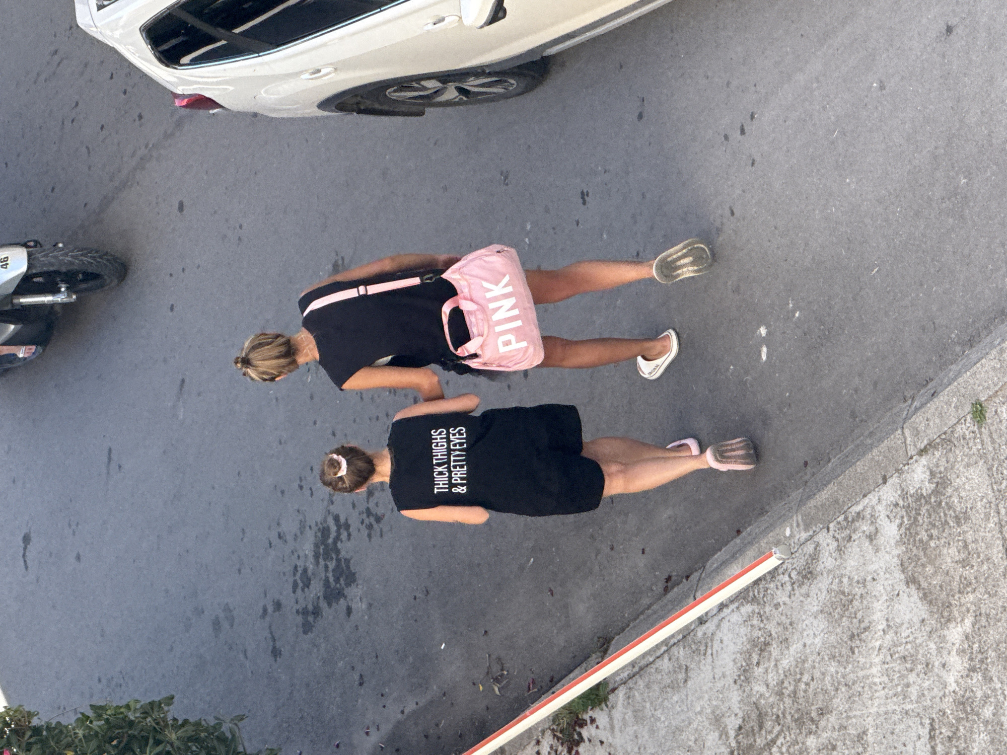 Two women walk side-by-side on a paved city street in Budva, Montenegro, with a white SUV parked to their right and a scooter in the background. The woman on the left wears a black sleeveless t-shirt with white text that reads "THICK THIGHS & PRETTY EYES" and black shorts, along with pink flip-flops. The woman on the right wears a black sleeveless top and black shorts, carrying a pink shoulder bag with "PINK" written in white letters. She has on white slides on her left foot and a grey sneaker on her right. The street appears to be an urban road with some wet patches and debris.