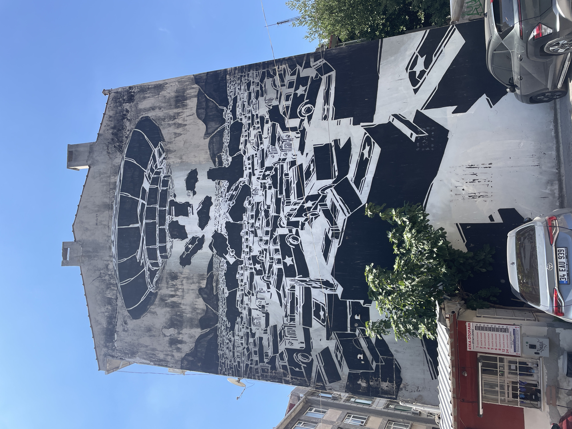 The photograph captures a large, black-and-white mural on the side of a tall, weathered building. The mural depicts a chaotic scene with numerous stylized cars, some appearing to be in a state of disarray or stacked upon each other. Dominating the top of the mural is a large, circular object resembling a UFO or a flying saucer, casting a shadow over the scene below. Mountains are visible in the background. To the left of the mural, part of an adjacent building is visible, as are trees and a small red structure with a sign displaying a price list. Two cars are parked in front of the mural. The sky is clear and blue. This mural is located on a building in the Moda neighborhood of Kadıköy, Istanbul, Turkey.