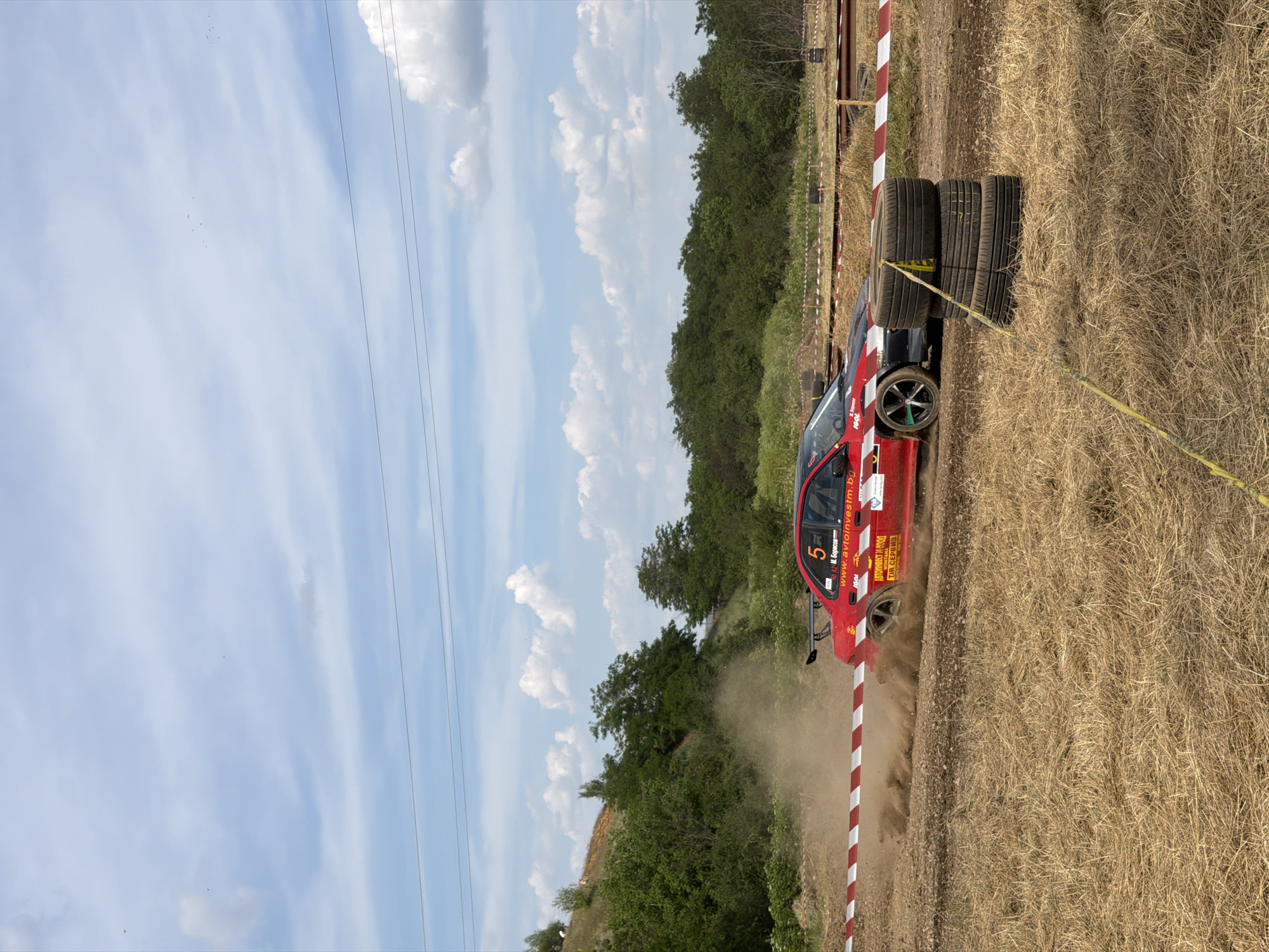 A red rally car with the number 5 on its side is captured in motion, kicking up a cloud of dust on a dirt track. The car, which appears to be a Subaru Impreza WRX, has a black hood and a prominent rear spoiler. It is positioned in the middle of the frame, turning to the right. In the foreground, there is a stack of three tires, bound together with yellow tape. The track is bordered by a red and white striped tape barrier. The background consists of dry, mowed grass, green trees, and a slightly hilly landscape under a bright blue sky with scattered white clouds. The scene suggests an outdoor motorsports event in a rural or semi-rural setting. The location is identified as Bahovitsa, Bulgaria.