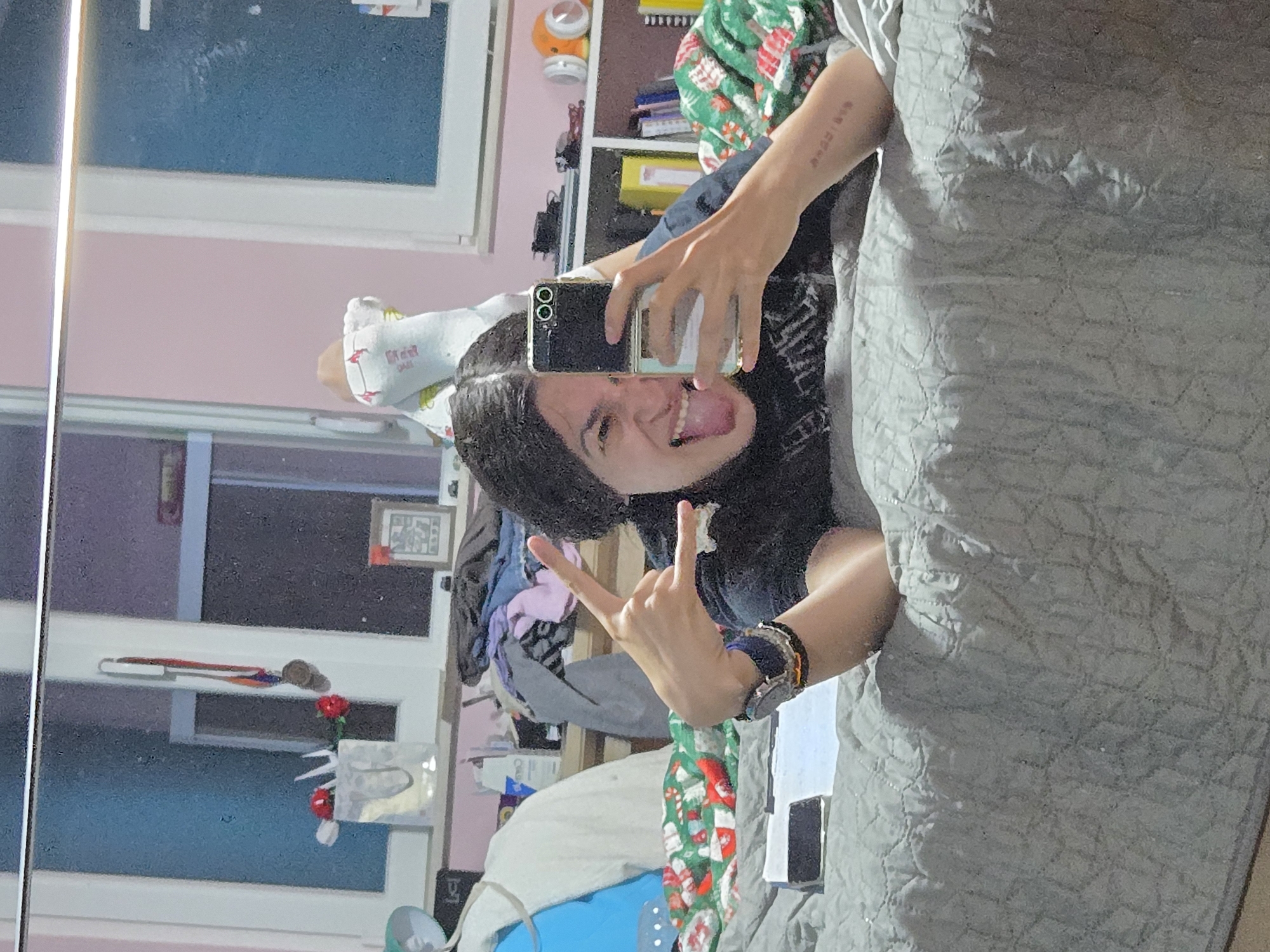 A person is visible in a mirror selfie. The person is sticking their tongue out and making a peace sign. The person is wearing a cast on one leg. A bedroom is reflected in the mirror. The location is not identifiable as Pleven, Bulgaria.