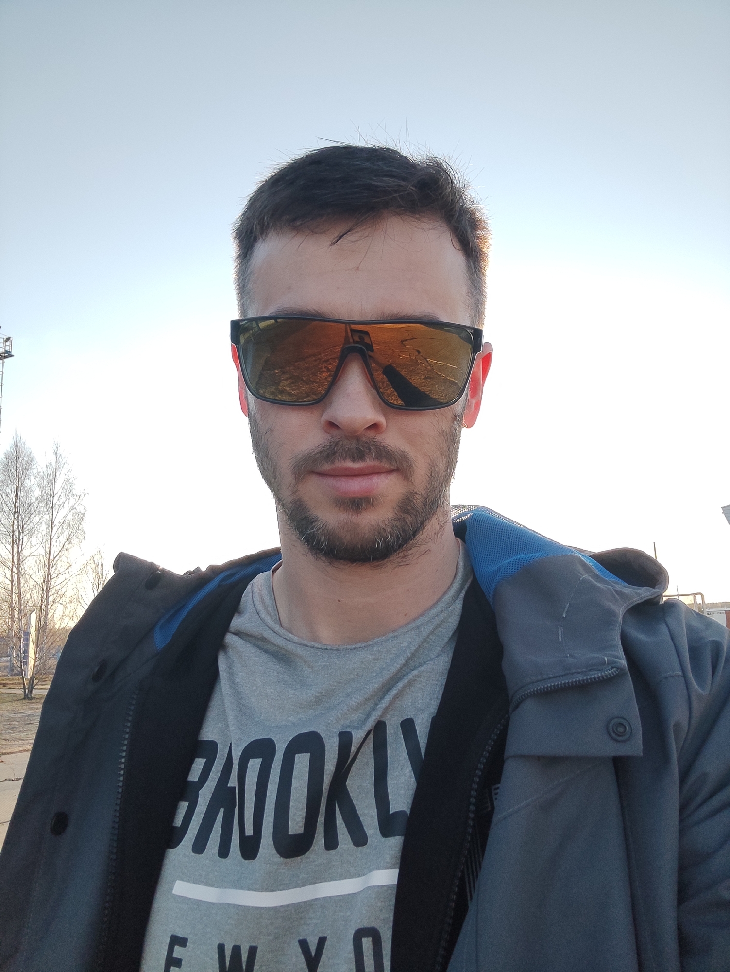A close-up, outdoor selfie features a man with short dark hair and a trimmed beard and mustache, looking directly at the camera. He wears dark, rectangular sunglasses with reflective orange-tinted lenses, which reflect a bright sky and possibly some distant ground or structures. His expression is neutral.
He is dressed in a light grey t-shirt that prominently displays the word "BROOKLYN" in dark capital letters across the chest, with "NEW YO" visible beneath it, suggesting "NEW YORK." Over the t-shirt, he wears a dark grey or charcoal jacket with a visible blue mesh lining around the neck and hood. The jacket has a zipper and snap fasteners.
The background shows a clear, bright, light blue sky, indicating fair weather. To the left, several bare-branched trees and a tall, dark industrial-like structure (possibly a crane or tower) are visible against the sky. The lighting suggests the photograph was taken during the golden hour, either late afternoon or early morning, with warm light highlighting the man's hair. The setting appears to be an open, possibly urban or semi-urban outdoor area.