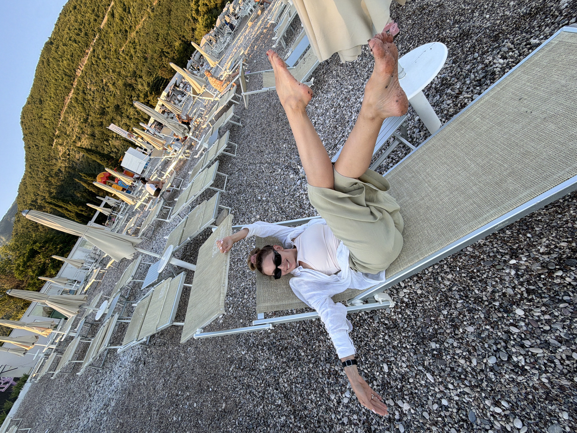 A woman with blonde hair pulled up in a bun, wearing sunglasses and a white open shirt over a pale pink tank top and olive green pants, is lying on a beige sun lounger on a pebble beach in Budva, Montenegro. She is posing with her legs extended and arms spread wide. The beach is lined with rows of empty sun loungers and closed umbrellas. In the background, a lush green mountainside rises, with a clear blue sky above. The sun is setting, casting a warm glow over the scene.