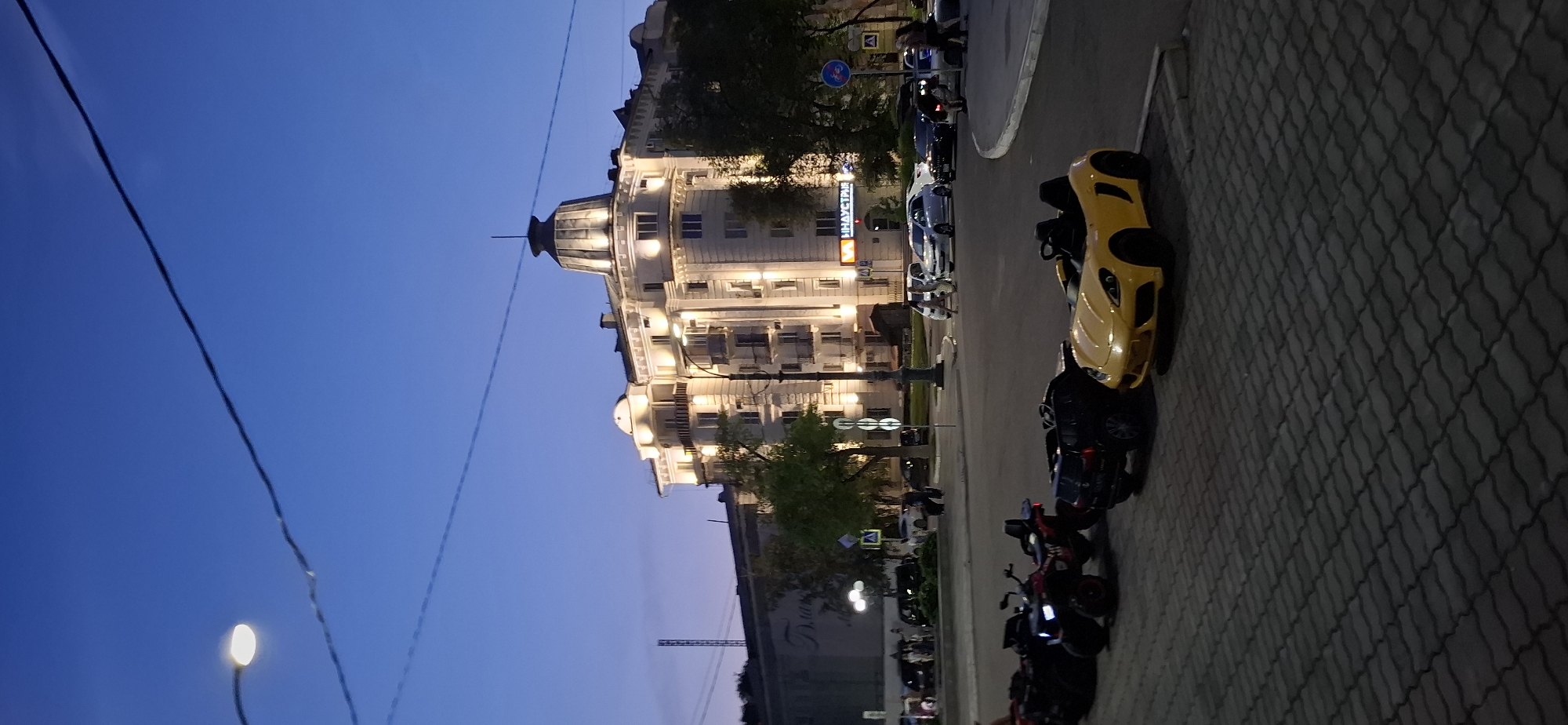 The image captures a street scene in Blagoveshchensk, Russia, at dusk. In the foreground, a line of children's electric ride-on vehicles, including ATVs and a yellow sports car, are parked on a paved area. Behind them, a street intersection is visible with several real cars, including a white sedan, a black SUV, and others. Pedestrians are walking in the vicinity. A large, brightly lit building with a domed roof and multiple balconies stands prominently in the background. The sky is a deep blue, indicating twilight. The overall impression is a public space transitioning from day to night.