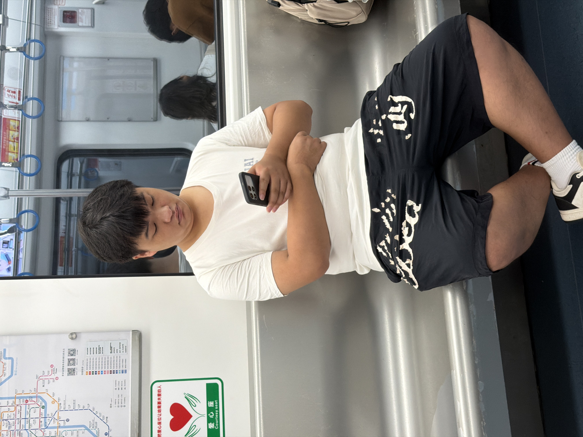 A young East Asian male is sitting on a metallic bench on a subway train, engrossed in his smartphone. He is wearing a white t-shirt and black shorts with white lettering. His posture is relaxed, with his arms crossed. In the background, other passengers are visible, some looking out the window or at their phones. A map of the subway system is displayed on the wall to the left, featuring Chinese characters and line indicators. The environment suggests an urban public transportation setting.