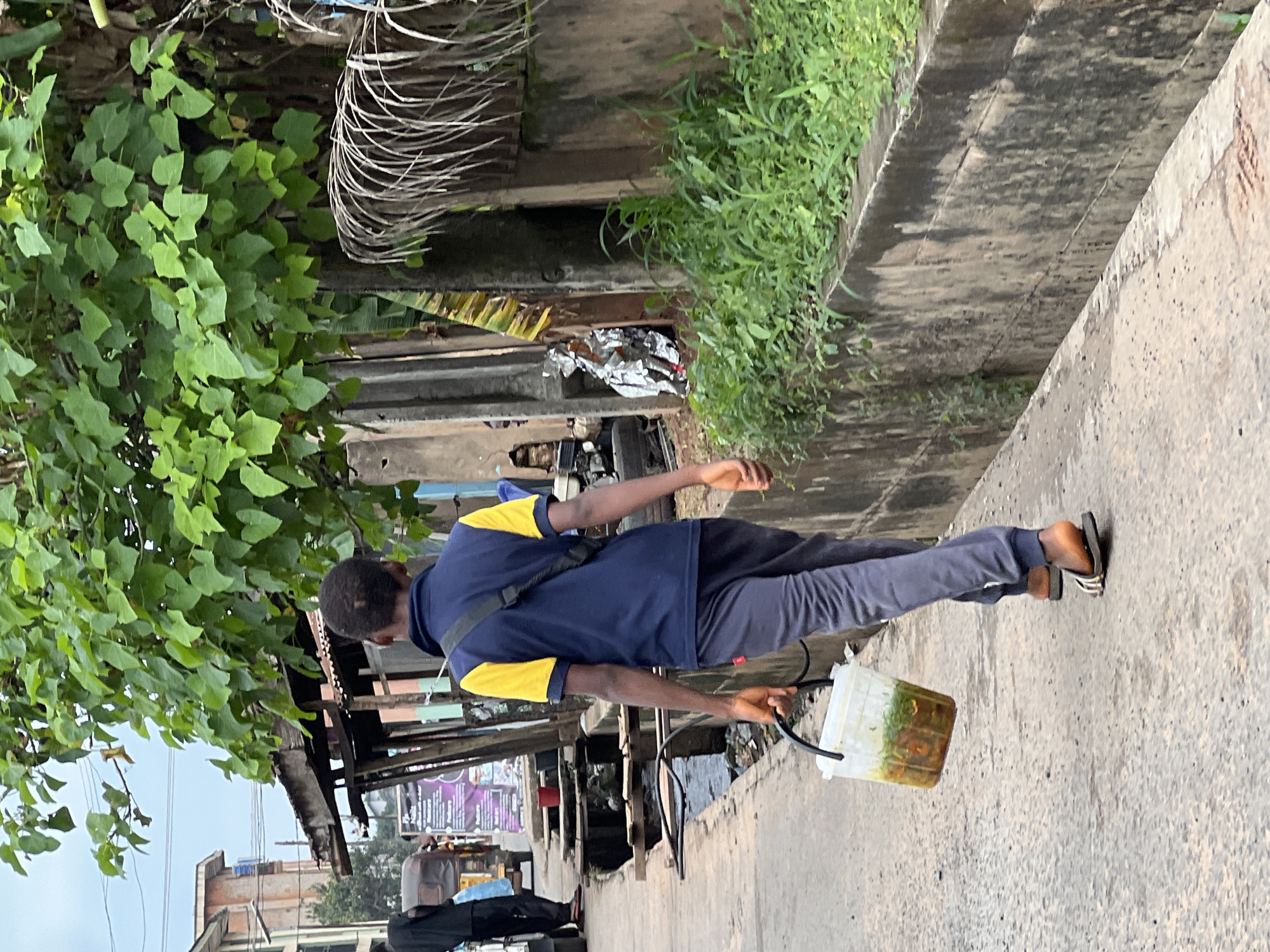 A person is walking on a concrete pathway next to a drainage ditch filled with murky water and debris. The person is wearing a blue and yellow shirt, grey pants, and sandals. They are carrying a translucent white container with a black handle, filled with a green and brown liquid. The pathway appears to be in an urban or semi-urban area, with buildings and vegetation visible in the background. The scene is dimly lit, suggesting it might be early morning or late afternoon. The overall impression is one of a mundane daily activity, possibly related to waste disposal or water collection, in a somewhat neglected environment. There is no visible text in the image.