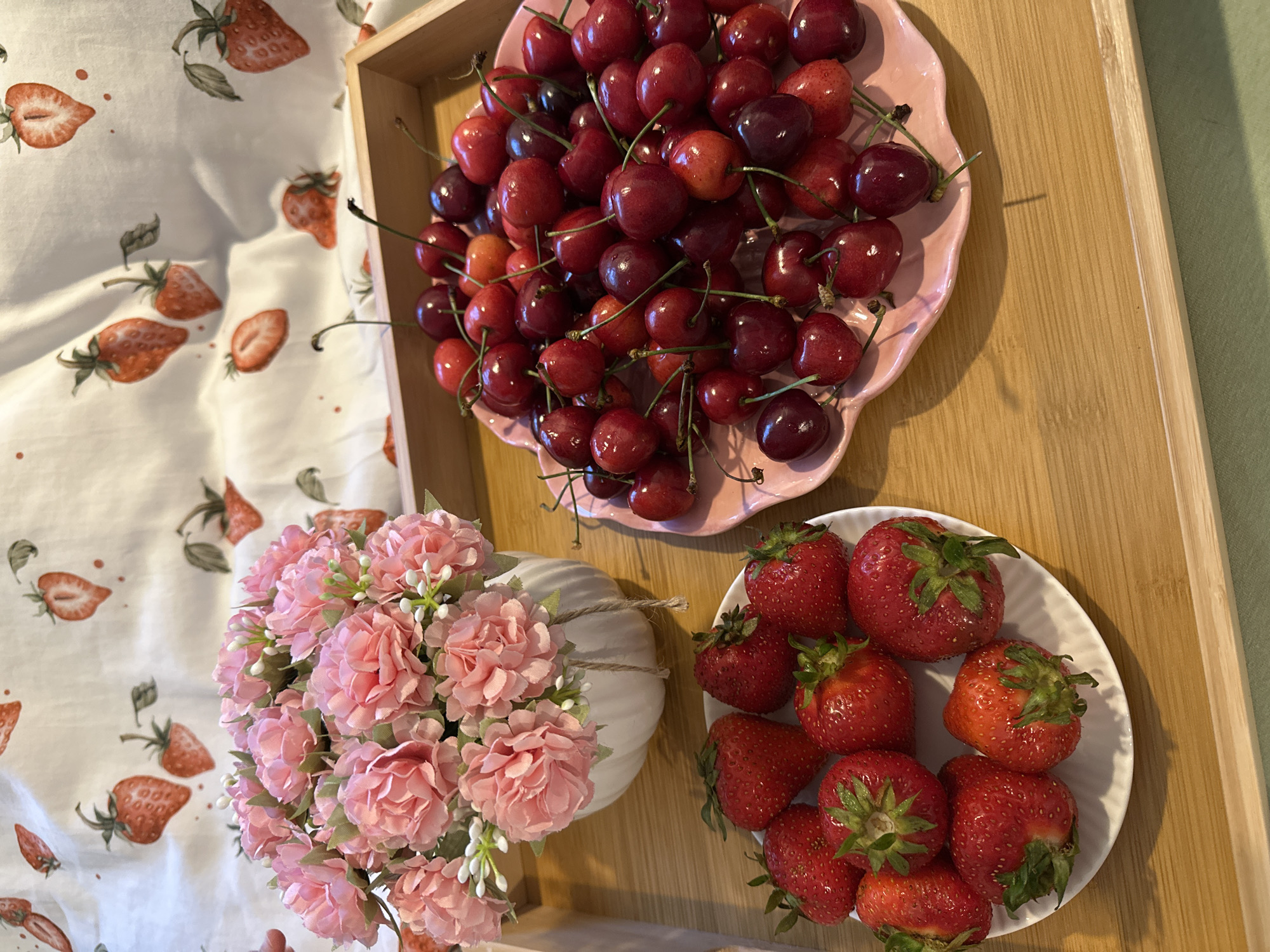 A wooden tray holds two plates of fruit and a vase of artificial flowers. On the left is a white plate filled with strawberries. On the right is a pink, heart-shaped plate piled with cherries. Behind the fruit is a white vase containing a bouquet of pink artificial flowers. The tray sits on a surface covered with a fabric patterned with strawberries. The image was taken in Wisła, Poland.