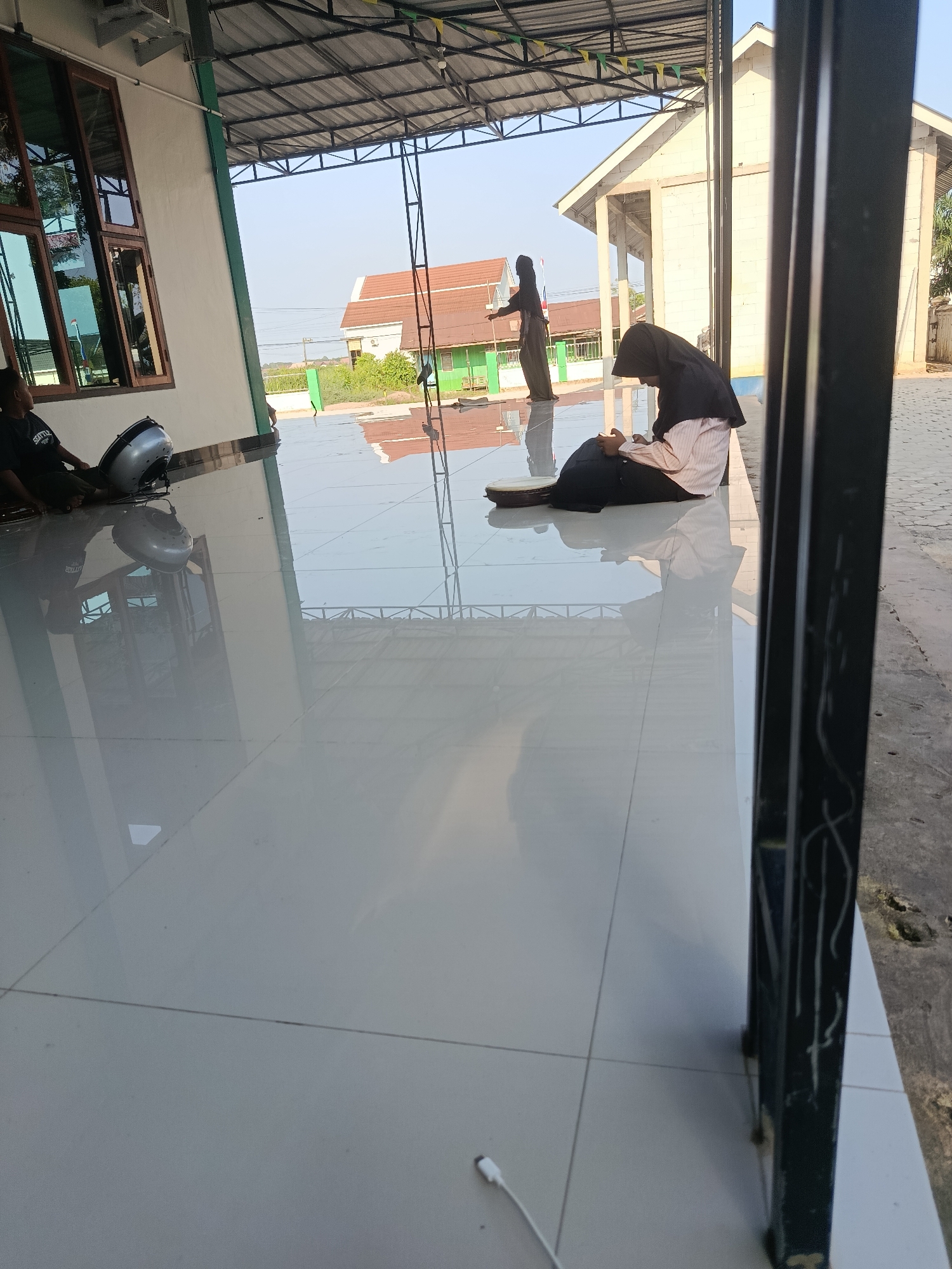 A low-angle, eye-level shot captures a tiled floor reflecting a covered outdoor area and a bright sky. In the foreground, a black metal pillar stands to the right. To the left, a person wearing a black t-shirt sits on the floor, leaning against a white wall with large windows. A silver fan rests on the floor beside them. In the middle ground, a person wearing a black hijab and a striped shirt sits on the floor, facing away from the camera, reading a book. Another person in a black hijab stands further back, facing left. Behind them, buildings with orange and white roofs and green walls are visible, set against a clear sky. The scene is outdoors, likely in a public or religious space, given the architectural style and the presence of individuals in religious attire. The location is identified as Swarga Bara, Indonesia.