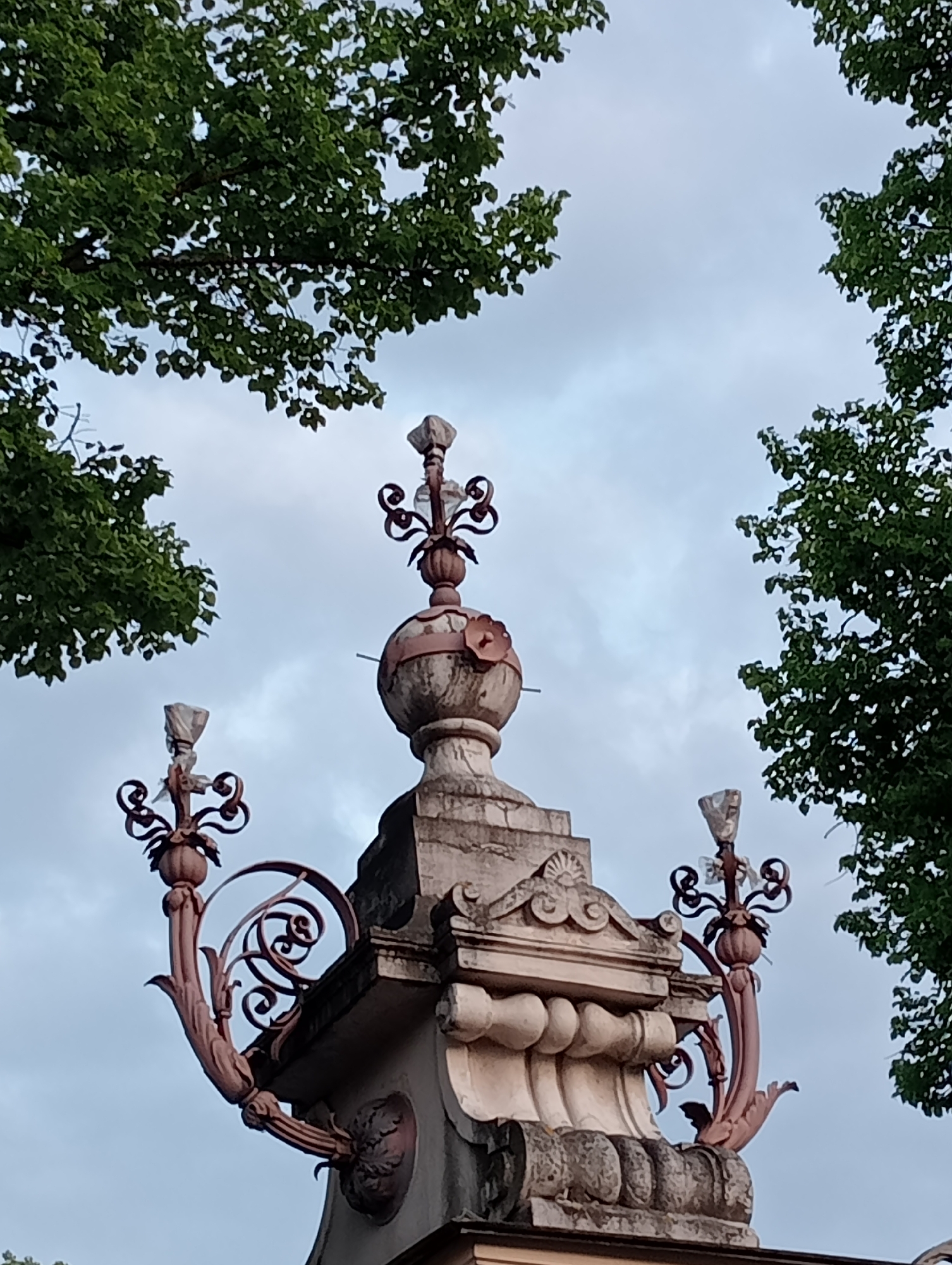 The image shows an ornate stone structure with decorative metal elements, possibly part of a fence or gate. The structure is topped with finials and features scrolling metalwork. Lush green trees frame the top of the image, and the sky is overcast. This appears to be a detail of the fence surrounding the Peter and Paul Fortress in Saint Petersburg, Russia, a historically significant site.