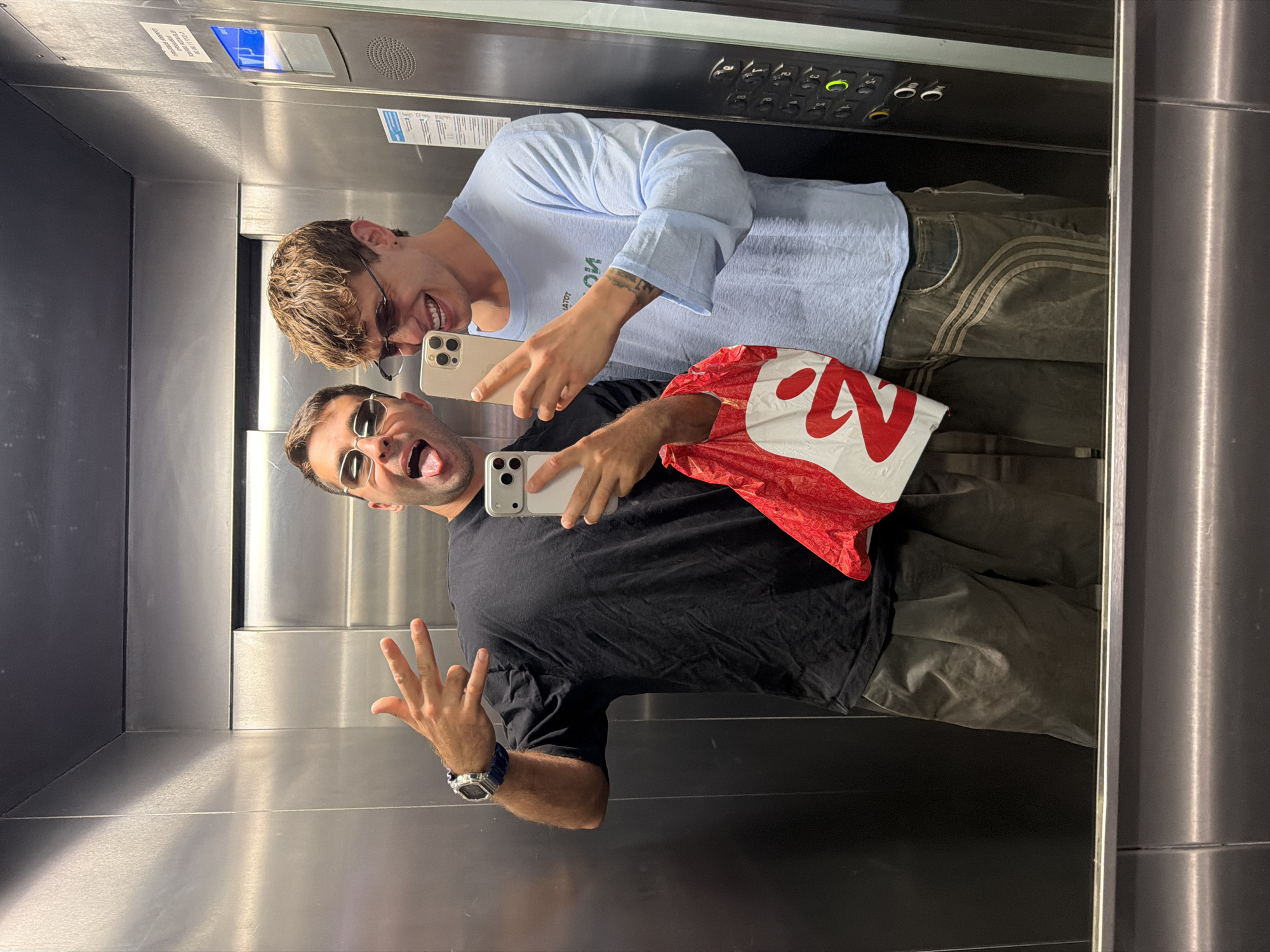 Two young men are taking a selfie in an elevator in Tbilisi, Georgia. The man on the left is wearing a black t-shirt, sunglasses, and dark pants. He is sticking his tongue out and making a peace sign with his left hand. He is holding a smartphone in his right hand. The man on the right is wearing a light blue long-sleeved shirt, sunglasses, and dark pants with stripes. He is smiling and holding a smartphone in his right hand. Between them, a red and white plastic shopping bag is visible. The elevator walls are made of stainless steel. The elevator buttons and a notice are visible on the right wall.