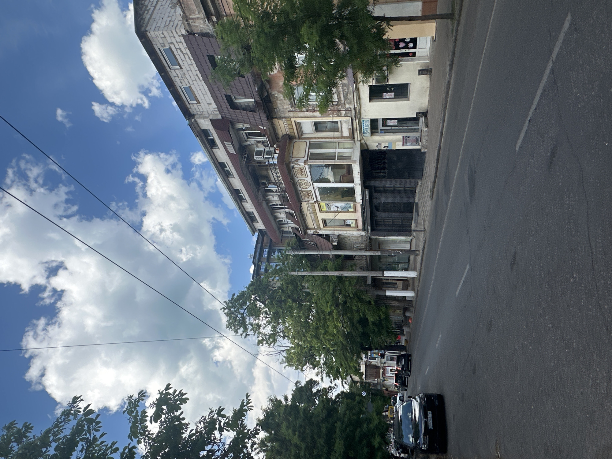 The image shows a street scene in Odesa, Ukraine, with a multi-story building on the left and a road on the right. The building appears to be old, with a mix of brick and plaster facades, and multiple windows and balconies. Trees and foliage are visible in front of and around the building. The road is paved and has lane markings. Several cars are parked or driving on the road. The sky is blue with scattered white clouds, suggesting it is daytime and the weather is fair. The perspective of the image is from ground level, looking down the street. There are some power lines visible in the sky. Some signs with text are visible on the building, but are not legible in detail.