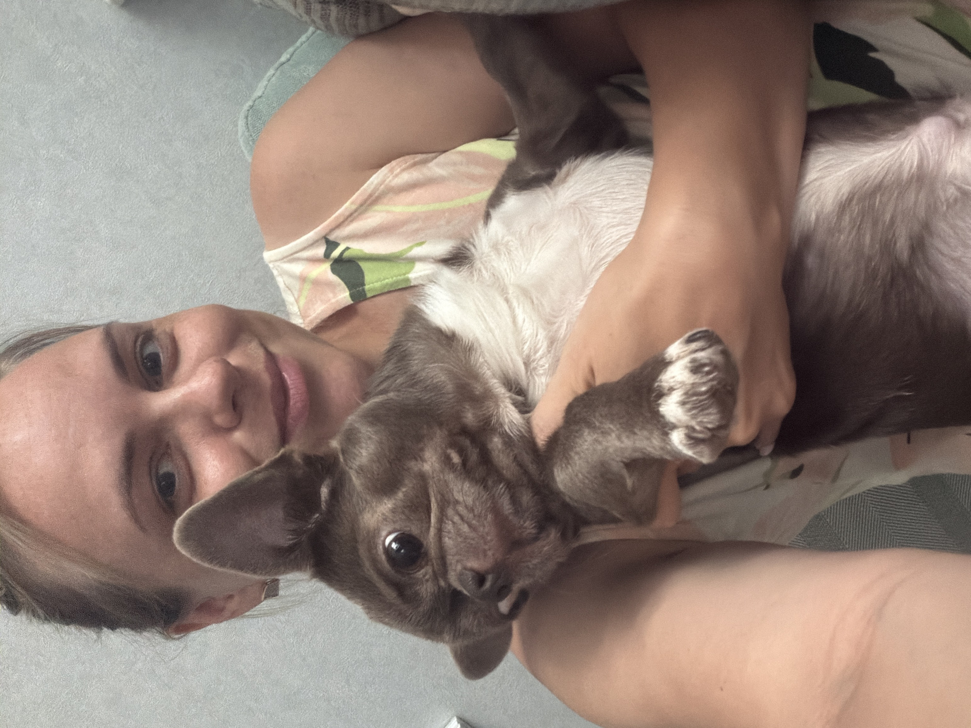 A blonde woman with her hair pulled back is holding a small brown and white dog. The woman is smiling at the camera and the dog is looking away from the camera. They are sitting on a sofa. The background is a light blue wall. The image was taken in Votkinsk, Russia.
