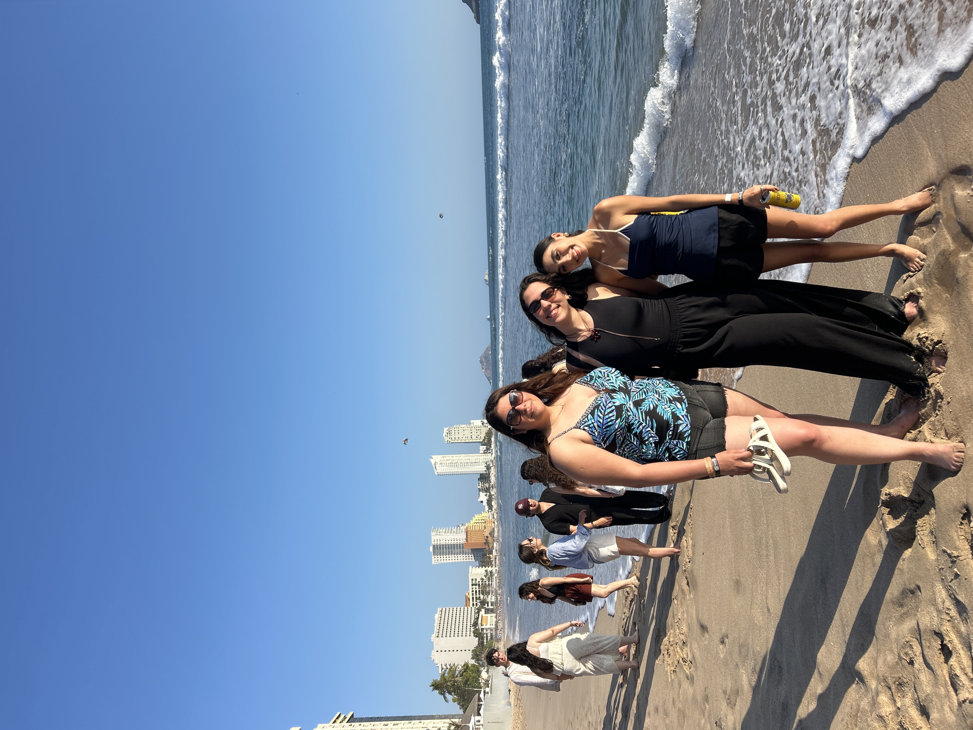 The image, taken on a bright, sunny day in Mazatlán, Mexico, captures a lively scene on a sandy beach. A group of people, predominantly young women, are gathered at the edge of the Pacific Ocean, posing cheerfully for a photograph.
In the foreground, four women stand in a line, smiling towards the camera. The leftmost woman wears a blue and green patterned top with black shorts, holding white sandals. Next to her, a woman in a black jumpsuit sports sunglasses. The third woman wears a navy halter top and black shorts, holding a yellow "PACIFICO" branded beer can. The fourth woman, on the far right, is dressed in a black top and black shorts. Their expressions are joyful and relaxed, reflecting the leisurely atmosphere.
Behind these primary subjects, several other individuals are visible further down the beach or partially in the water. One person wears a black top and a maroon baseball cap featuring an "NY" logo.
The setting consists of a wide, sun-drenched sandy beach with gentle ocean waves lapping at the shore. The water is a clear blue, and the sky above is a vast, cloudless expanse of bright blue, with a small parasailer or kite visible in the distance. Along the coastline, a modern city skyline with numerous tall, light-colored buildings extends into the background, signifying a vibrant coastal resort area. The bright sunlight casts distinct shadows on the sand, indicating it is likely midday or early afternoon.