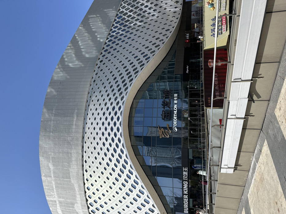 A modern building with a curved facade featuring a decorative, perforated white outer layer. Below, large glass windows showcase the storefronts for Burger King and Decathlon. Signs in both English and Chinese are prominently displayed. Other brands and establishments are visible on signs above, including Pizza Hut. Street-level architectural elements are visible, along with a bright blue sky in the background.