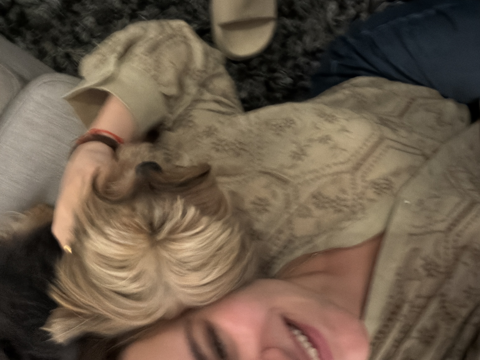 A blurry, high-angle image shows a person lying down, possibly on a sofa or bed, with a small dog resting its head on the person's shoulder. The person is wearing a light-colored, patterned shirt and blue jeans. A sandal is visible near the top of the frame. The background includes a gray rug and a light-colored cushion or part of the sofa. The person is smiling slightly. No identifiable location in San Bernardino Tlaxcalancingo, Mexico can be determined from the image.