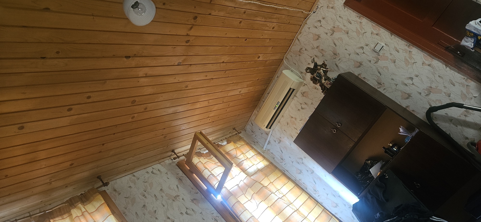 The image depicts an interior room with a wooden slatted ceiling and walls. On the wall, there is an air conditioning unit and patterned wallpaper. A dark wooden cabinet with two doors is visible, with items on its shelves and a baby stroller next to it. To the left, a window is covered by yellow and orange checkered curtains. A light fixture is attached to the ceiling. The scene appears to be a domestic living space. The location is identified as Lyaskovets, Bulgaria.