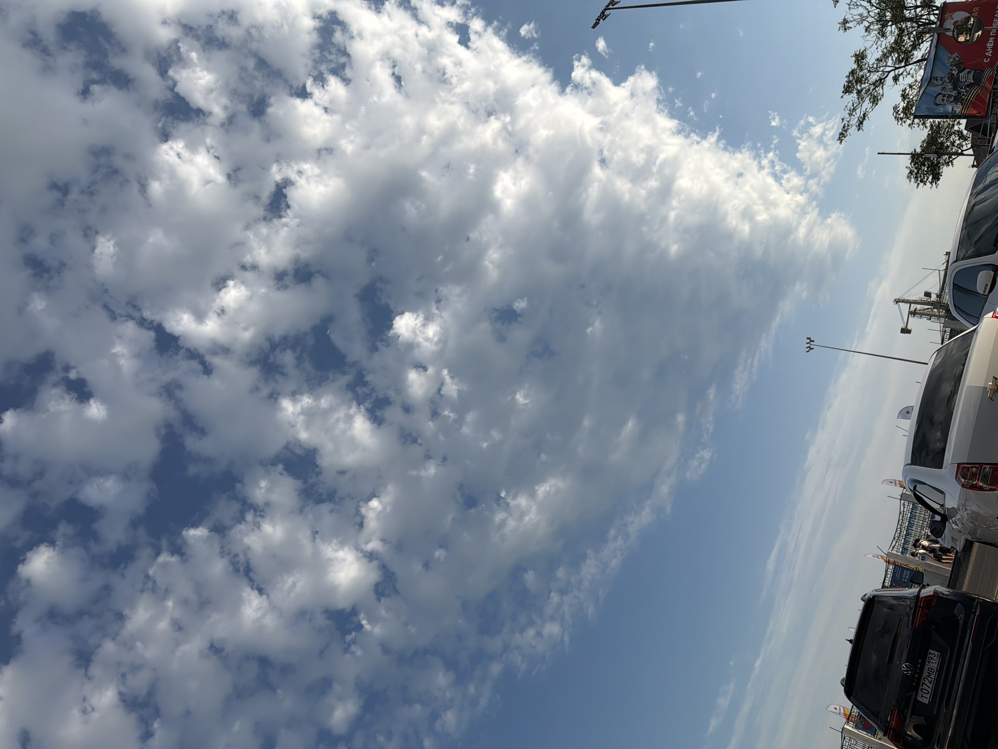 The image depicts a sky filled with a dramatic formation of white, puffy cumulus clouds against a bright blue sky. In the foreground, several cars are visible, suggesting a street or parking lot setting. A black Volkswagen Tiguan with a license plate reading "T072MB 193" is parked on the left. A white Chevrolet Cobalt is parked to its right.
Behind the cars, a fence with several flags can be seen. Further in the background, construction cranes are visible, indicating ongoing development or infrastructure work. A large poster featuring portraits of people is also present on the right side of the image. The overall impression is a clear day with a visually striking sky, possibly in an urban or industrial area. The location is identified as Tuapse, Russia.