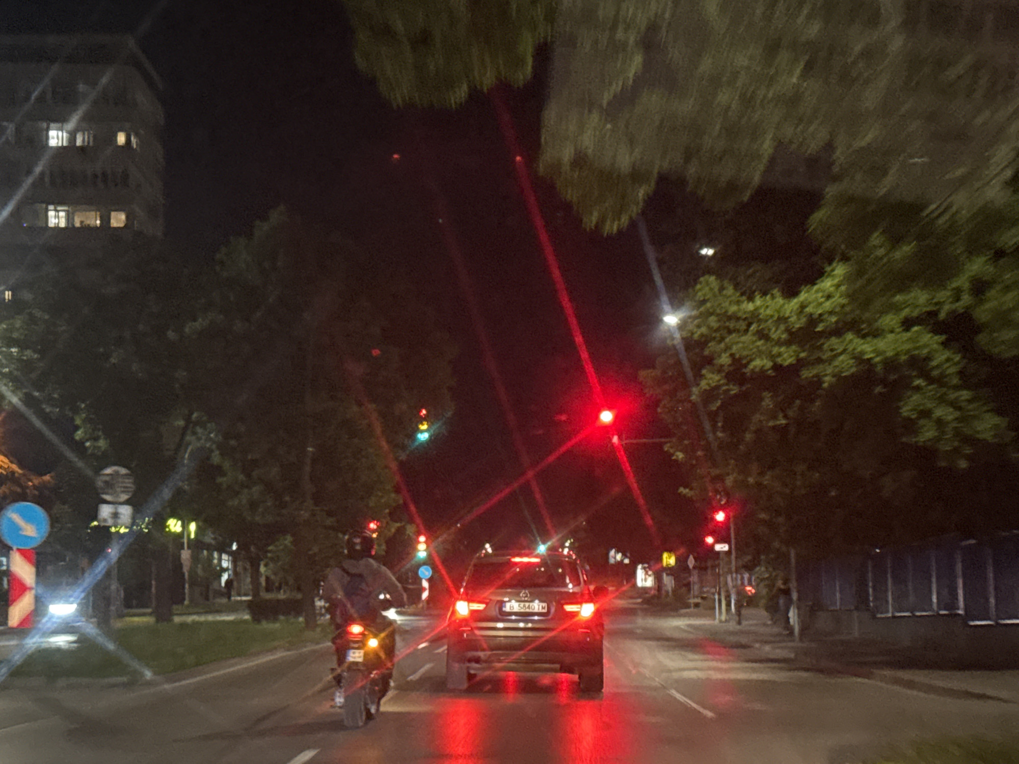 The image depicts a nighttime view from a vehicle on a city street in Varna, Bulgaria. The scene is illuminated by streetlights, vehicle lights, and traffic signals.
In the foreground, a motorcycle is visible on the right lane, with its brake lights illuminated. The rider is wearing a helmet. Ahead of the motorcycle, a black SUV is in the same lane, with its brake lights on, indicating it is stopped or slowing down. The license plate of the SUV is partially visible as "B 5840 TM".
Further ahead, traffic lights are visible, with the topmost lights illuminated red, suggesting traffic is stopped. Trees line the sides of the road, with some branches overhanging the street, creating a canopy. On the left side of the road, a tall building with illuminated windows is visible in the background. Road signs, including a blue circular sign with a white arrow and a white and red striped barrier sign, are present on the left. The road surface appears wet, reflecting the lights. The overall impression is that of a common urban scene at night, potentially at an intersection.