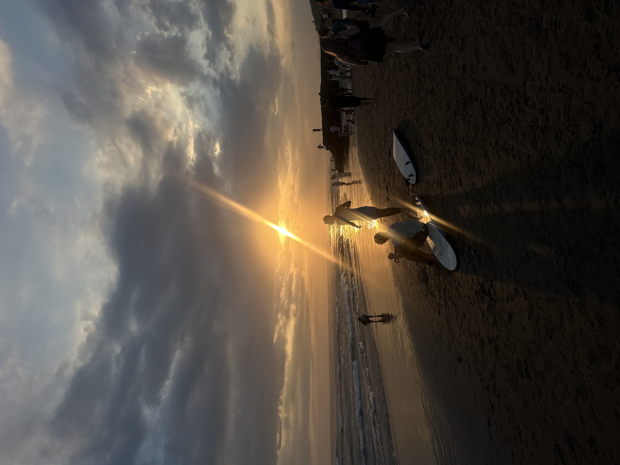 The image depicts a beach scene at sunset, with the sun casting a bright, lens-flare effect across the water and sky. Dark clouds are scattered across the sky, contrasting with the warm glow of the setting sun. The ocean is visible with gentle waves rolling onto the shore, creating a wet, reflective sand surface. Several people are scattered along the beach, some walking, some standing, and two are seated in the foreground. Two surfboards are lying on the sand near the seated individuals. The overall impression is one of a relaxed, end-of-day gathering on a beach.