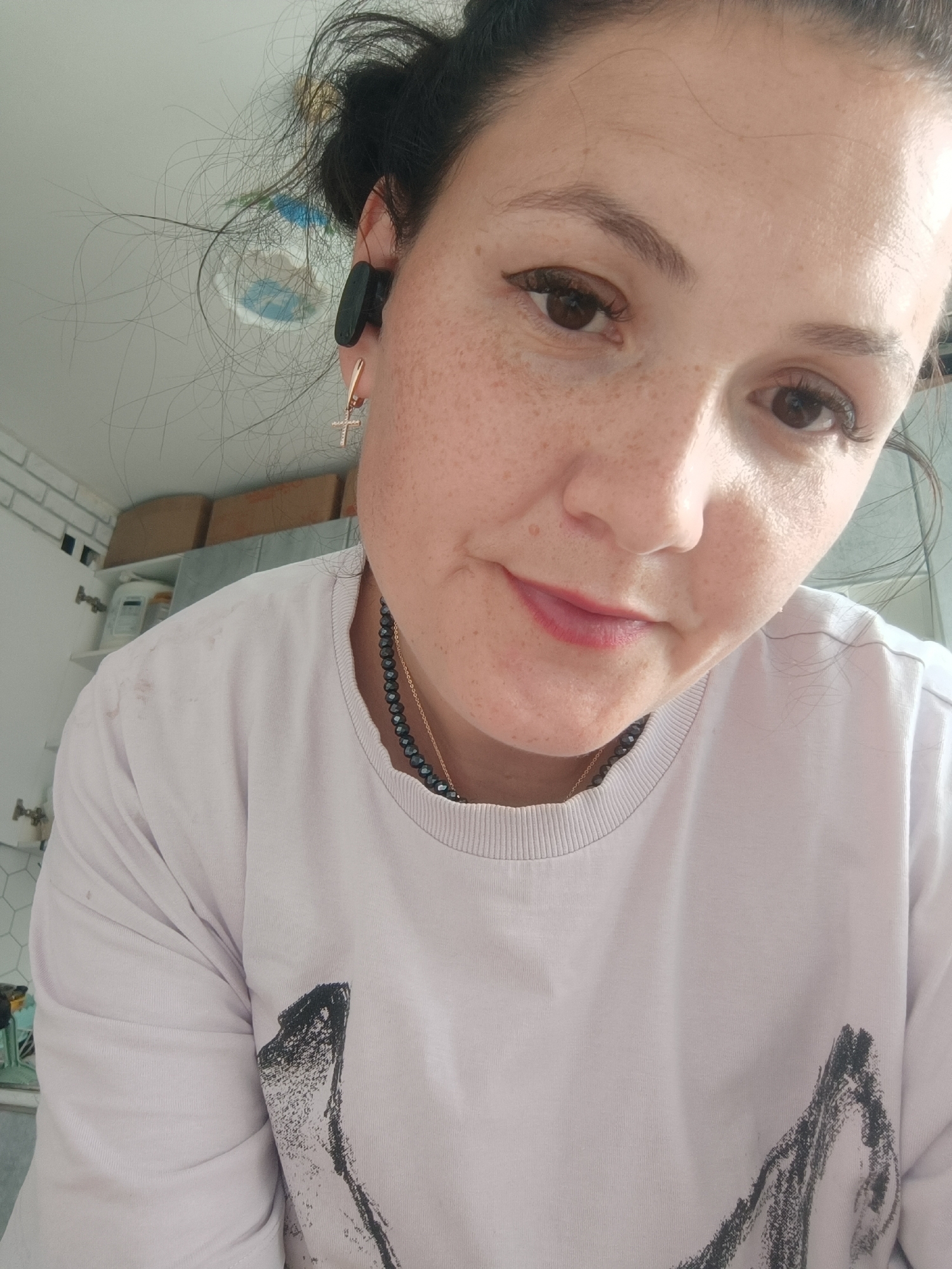 A close-up, eye-level, slightly high-angle, outdoor selfie shows a woman in her late 20s or early 30s with fair skin, dark hair pulled back loosely, and freckles across her nose and cheeks. She is wearing a light purple sweatshirt with a black abstract design on the chest. She has dark eyeliner and mascara, and her lips are painted a soft pink. A dark bead necklace with a gold chain is visible around her neck, and a small gold cross earring hangs from her right ear. A black earbud is in her right ear. The background is blurred, but hints of shelves with boxes and bottles suggest an indoor setting, possibly a bathroom or storage area, in Chaikovsky, Russia. The lighting is natural and appears to be coming from above.