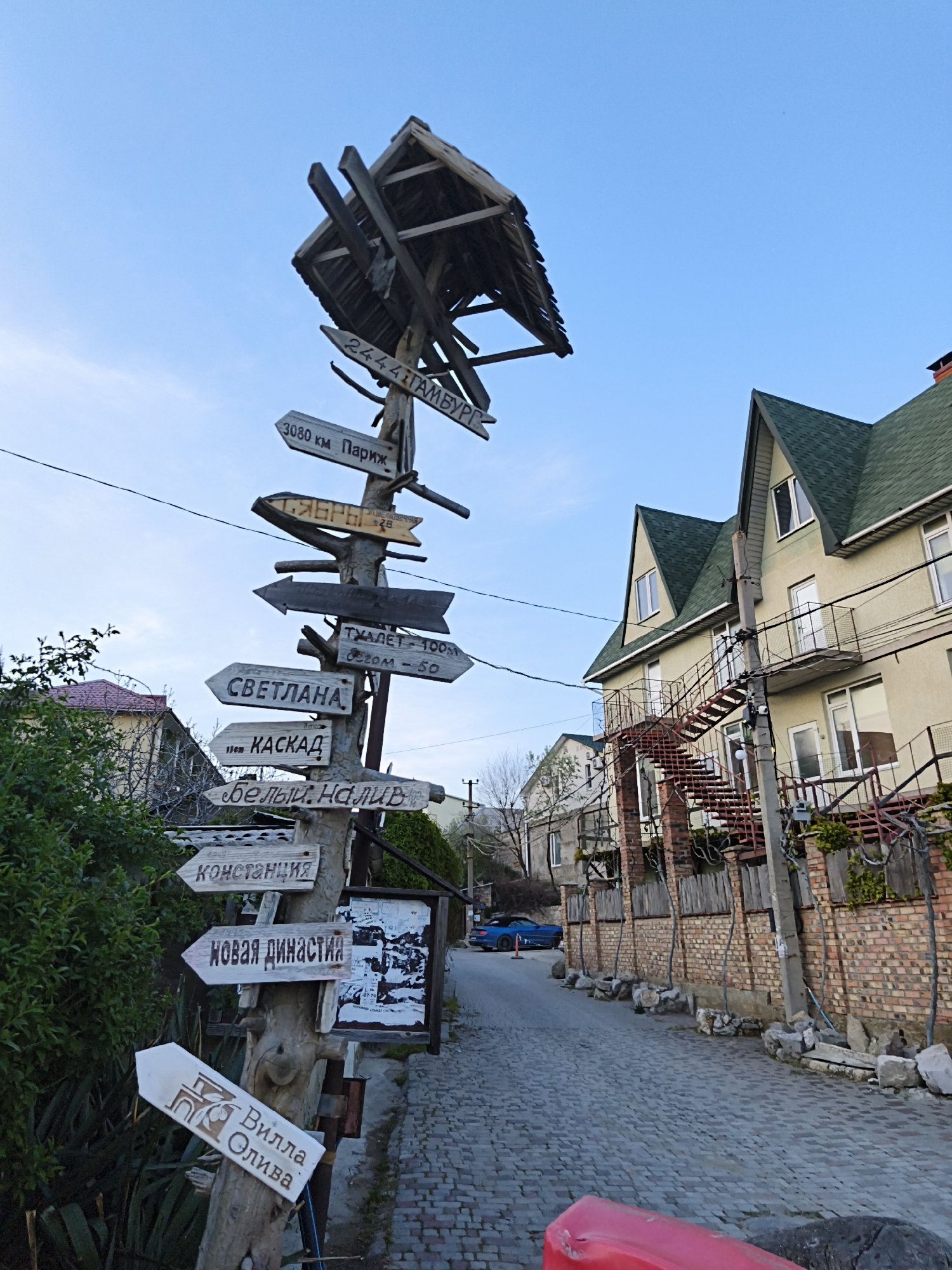 The image depicts a weathered signpost in Орджоникидзевский поселковый совет, Ukraine, adorned with multiple directional signs indicating various destinations, including "Гамбург," "Париж," "Светлана," and a "Вилла Олива." The signpost, crafted from a tree trunk, features a rudimentary wooden shelter at its peak. The immediate surroundings consist of a narrow, cobblestone street flanked by buildings, one of which is a multi-story structure with a green roof and an external metal staircase. The scene is captured under a clear, blue sky. A glimpse of a blue car and scattered vegetation contribute to the urban setting.