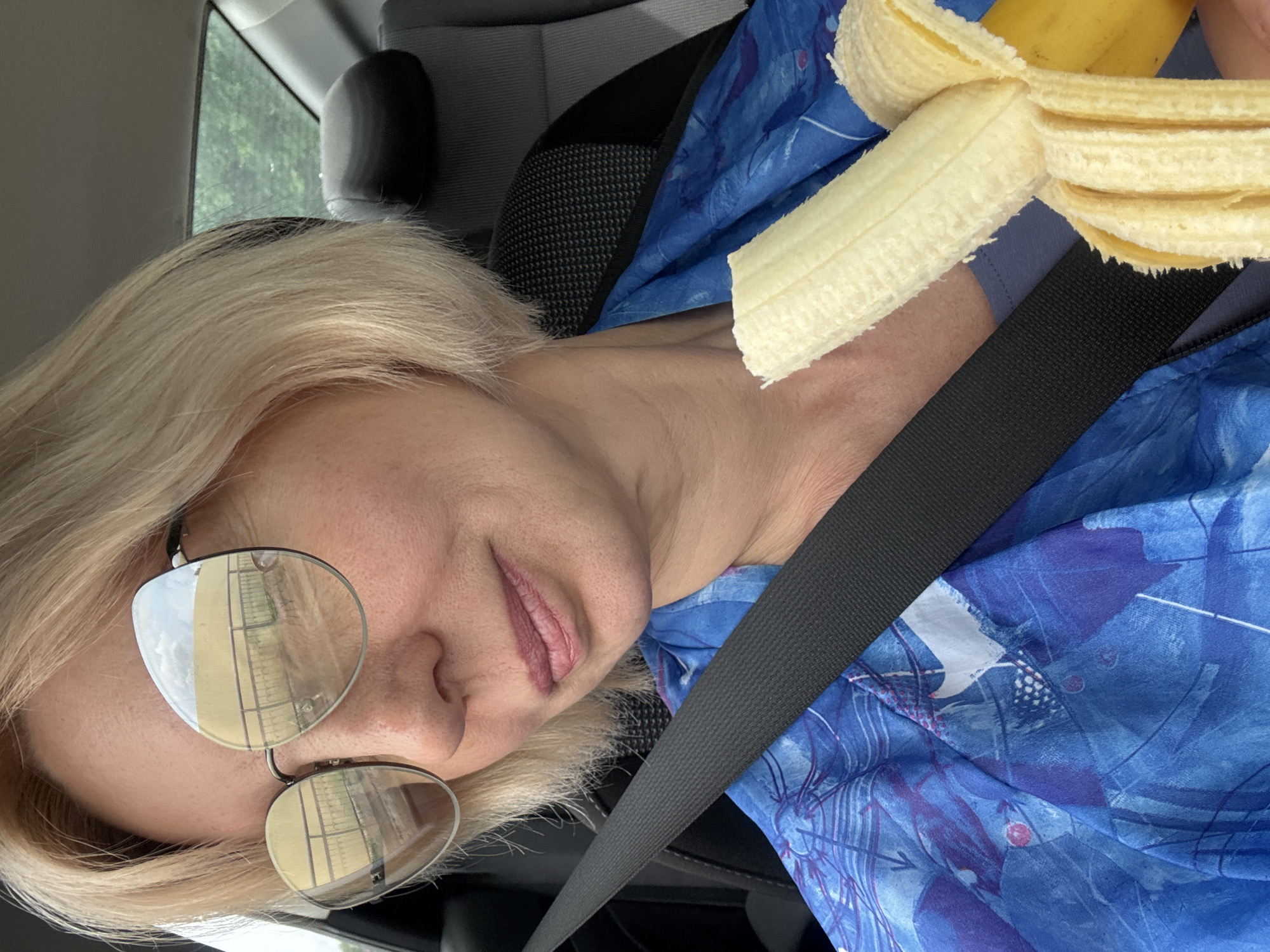 A woman with short blonde hair, wearing large, round sunglasses and a blue, abstract-patterned shirt, is seated in the passenger seat of a car. She is holding a partially peeled banana in her right hand, bringing it towards her mouth. Her left hand is not visible. A black seatbelt is visible across her chest. The background of the car interior, including a dark headrest and a window showing greenery outside, is visible. The image does not contain any information about the city of Teshebs, Russia.
