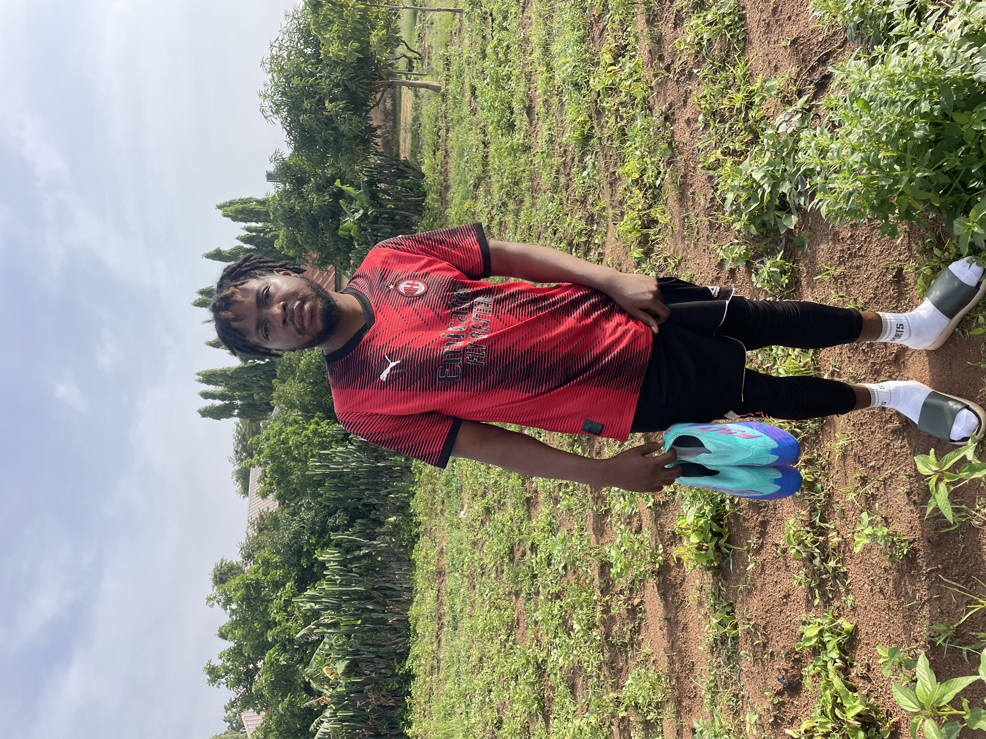 A young Black man stands in a field of green plants and dirt under a cloudy sky. He is wearing a red and black striped short-sleeved t-shirt, black shorts, black leggings, white socks, and dark green slides. He holds a pair of turquoise and purple cleats in his right hand. In the background, there are trees and a building with a red roof. The location is Gura Top, Nigeria.