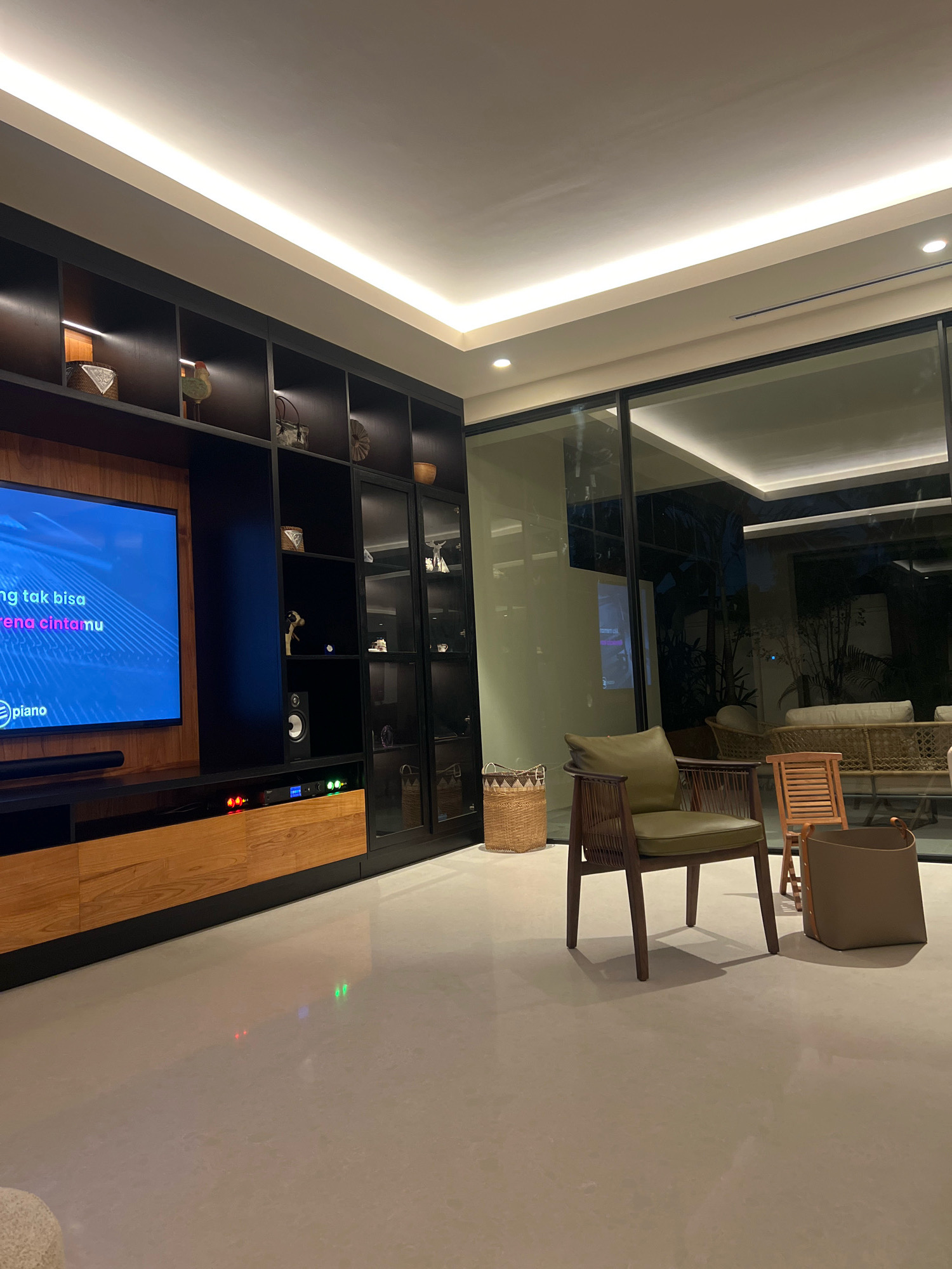 This is a wide-angle, eye-level shot of a modern living room in Ngaglik, Indonesia. The room is illuminated by recessed ceiling lights and accent lighting within a large built-in entertainment unit. The entertainment unit, made of dark wood and glass, dominates the left side of the frame, housing a large television screen displaying what appears to be karaoke lyrics. Below the TV is a wooden cabinet with visible electronics, some with colored indicator lights.
To the right of the entertainment unit, a spacious glass wall offers a view into an outdoor area with lush greenery and outdoor furniture, suggesting a connection between the indoor and outdoor spaces. In the foreground, a minimalist aesthetic is evident with a light-colored, polished floor. Positioned in the center-right of the room are several pieces of furniture: a dark wood armchair with a green cushioned seat, a small wooden side table, and a taupe tote bag resting on the floor. A woven basket is placed behind the armchair. The overall impression is one of contemporary design and comfortable living.