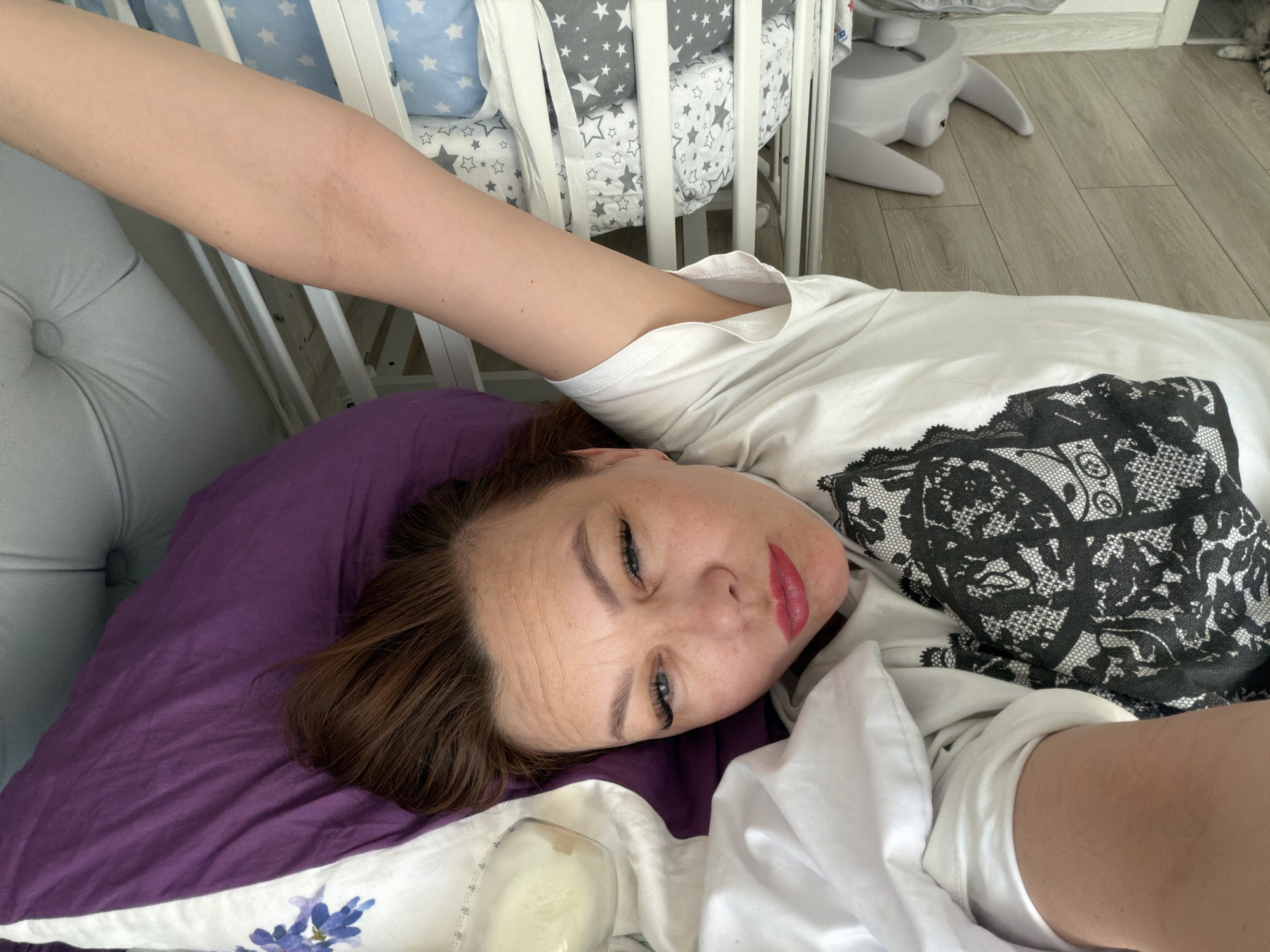 A female adult subject is depicted lying supine on a bed or similar surface, positioned with her head on a purple pillow. Her head is angled right, facing the camera with partially open blue eyes, prominent eyelashes, defined eyebrows, and red lipstick. Her hair is reddish-brown. She wears a white short-sleeved top featuring a black lace-patterned design across the chest. Her right arm is extended upwards and out of frame; her left arm is partially visible.
The immediate surroundings include a light gray tufted upholstered object to the left. Behind the subject's head, a white crib railing is visible, containing star-patterned bedding (white stars on blue, white stars on gray). A light wood-planked floor is visible beneath the crib and extending to the right, where a white, multi-legged infant bouncer is positioned. A portion of a striped feline is visible in the upper right background. The environment is an interior space, brightly illuminated. This scene is located in Chaikovsky, Russia.