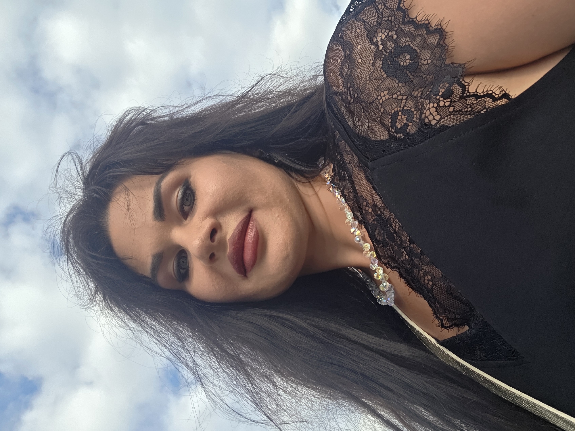 This media file shows a close-up, Dutch angle shot of a woman outdoors against a cloudy sky, likely in Sochi, Russia. The woman has long, dark hair and is wearing a black top with black lace detailing on the shoulder and neckline, and a necklace made of clear and iridescent beads. Her makeup includes dark eyeliner and lipstick in a deep berry shade. She is looking towards the camera with a subtle smile, her expression appearing confident and perhaps a little playful. The lighting suggests it is daytime, and the scattered clouds indicate fair weather. The low angle of the shot, looking up at the woman and the sky, gives a sense of her dominance or importance in the frame. There is no visible text in the image.