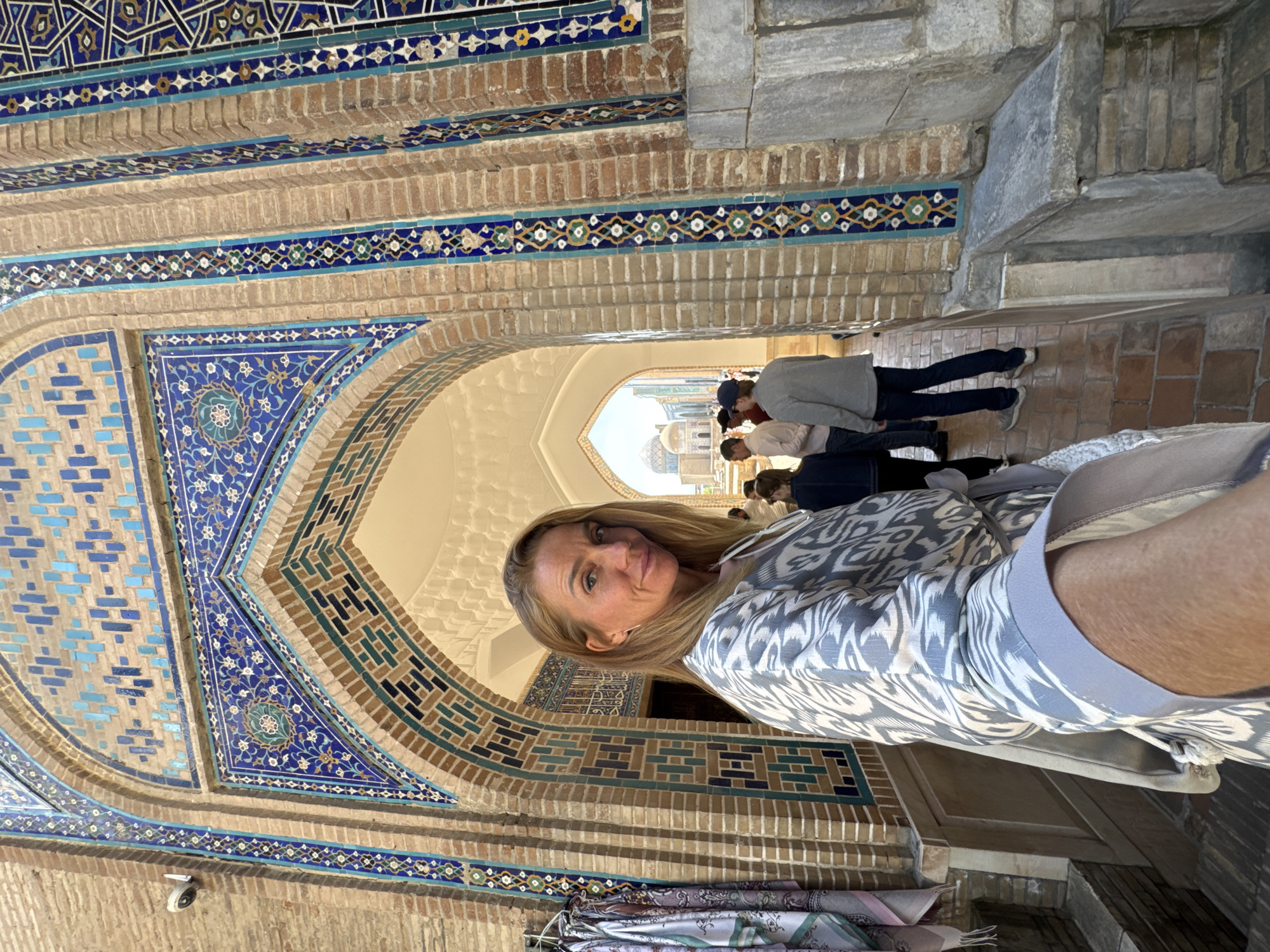 The image shows a woman posing for a selfie inside or in front of a structure adorned with intricate tilework. The tilework features geometric patterns and floral designs in shades of blue, brown, and white, and there are also people in the background walking through an archway. A dome is visible further back in the structure. Based on the architecture and design, it is highly probable that the location is in the city of Samarkand, Uzbekistan, which is known for its historical sites and Islamic architecture.