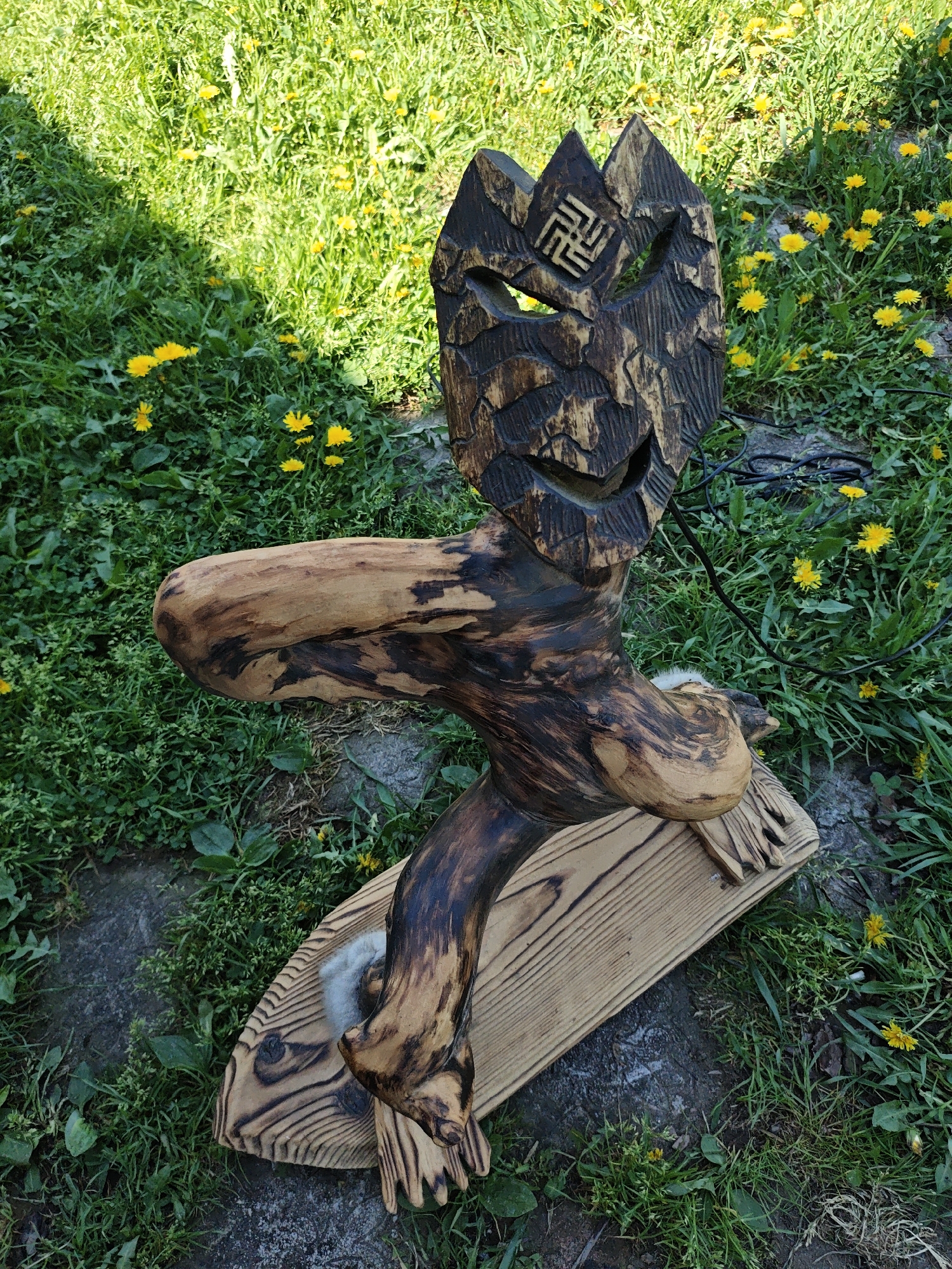 The image depicts a carved wooden sculpture of a humanoid figure in an outdoor setting, likely in Боровково, Russia. The sculpture is placed on a wooden base and positioned amongst grass and dandelions. The figure's head features a decorative pattern reminiscent of mountains and an emblem with a geometric design etched into the wood. The figure's body appears to be crafted from tree limbs and has been stained or burned to create a color contrast. There is also some material, possibly white fur or cotton, affixed to the sculpture near its midsection and the base. A black cable or wire is visible behind the sculpture. The ground is covered in grass interspersed with yellow dandelions. The sculpture is partially in shadow.