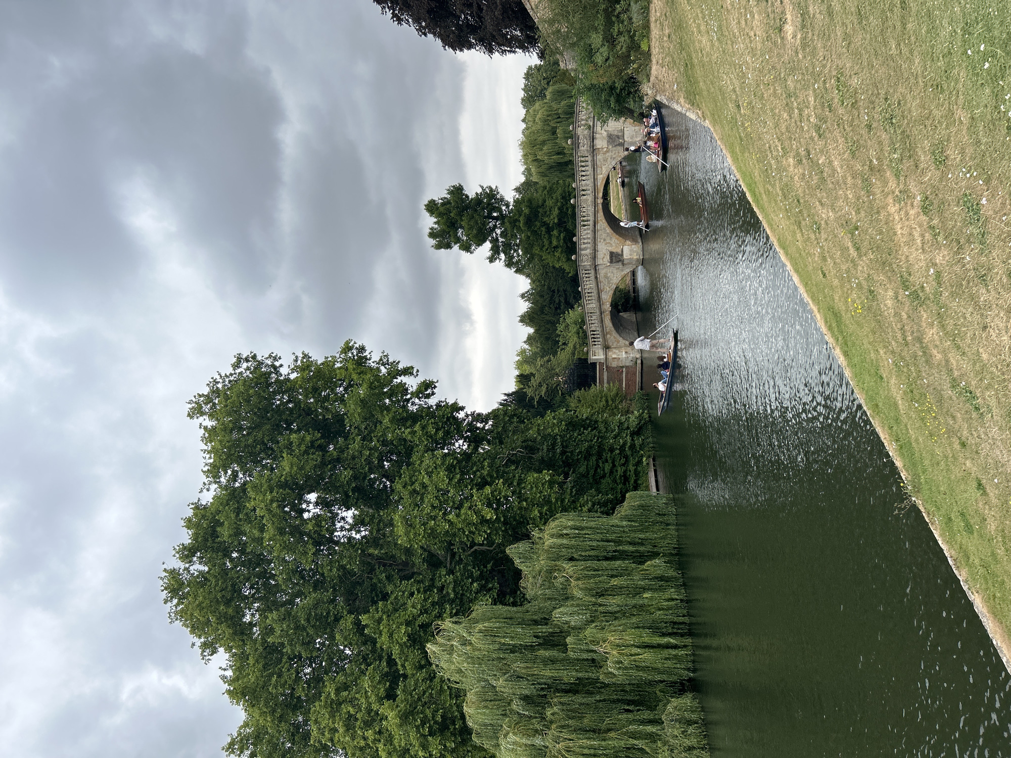 The image depicts a serene scene on the River Cam in Cambridge, United Kingdom, under a cloudy sky. A stone bridge with multiple arches spans the river, with several punts, traditional flat-bottomed boats, navigating beneath it. On the punts, individuals are engaged in punting, a leisurely activity where one propels the boat using a long pole. The riverbanks are lined with lush greenery, featuring mature trees, including a prominent weeping willow on the left, and manicured lawns. The overall atmosphere is peaceful, with a sense of calm recreational activity on the water.