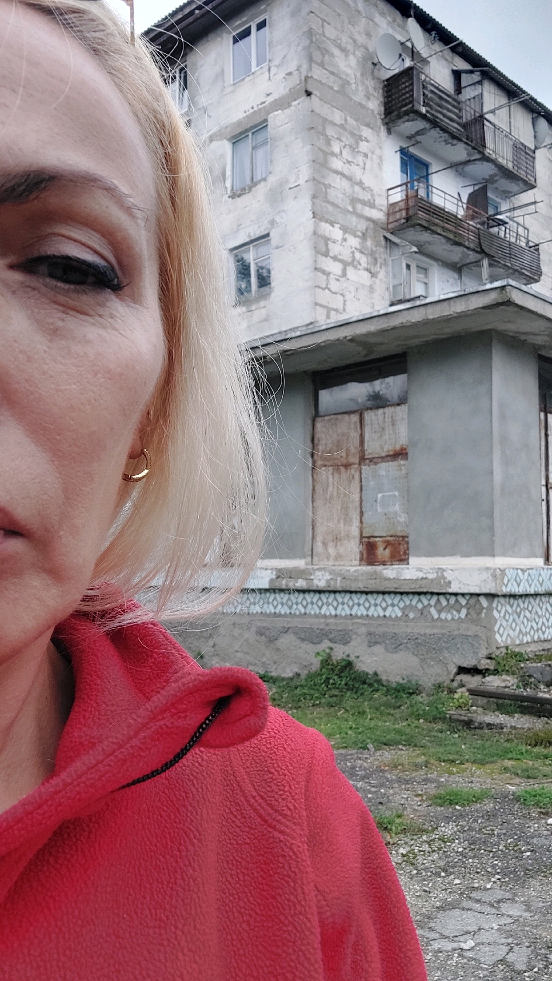 The image shows a close-up of a woman with blonde hair and makeup, wearing a red fleece jacket. She is standing outdoors in front of a weathered, concrete apartment building. The building appears to be in disrepair, with peeling paint and rusted balconies. The area around the building is overgrown with grass and gravel. The woman's face is partially visible, and she is looking slightly to the left. The background suggests an urban or semi-urban environment in Frunză, Moldova, characterized by older, concrete structures.