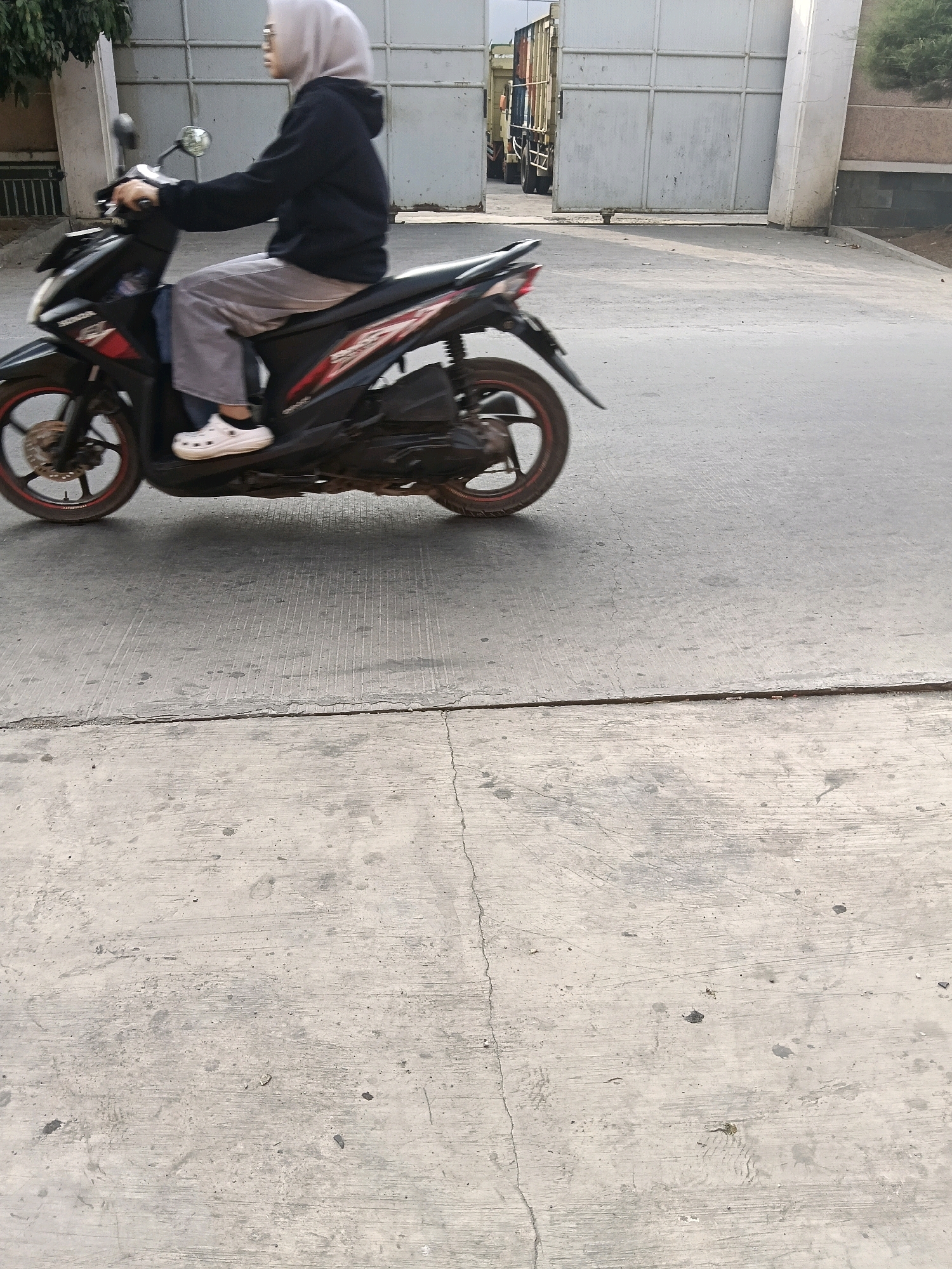 A person wearing a grey hijab and dark hooded jacket is riding a black and red motorcycle on a paved road. The person is wearing grey pants and white Crocs shoes. The motorcycle has a red stripe along its side and a black rear fender. In the background, there is a large metal gate, and a truck is partially visible through the gate. The scene appears to be in an industrial or commercial area, possibly a warehouse district, given the presence of the gate and truck. The road is concrete and appears to be in good condition, with some tire marks visible. The lighting suggests it is daytime. The image does not provide specific information to pinpoint the exact location within Belimbing, Indonesia, but the context implies an urban or semi-urban industrial setting.