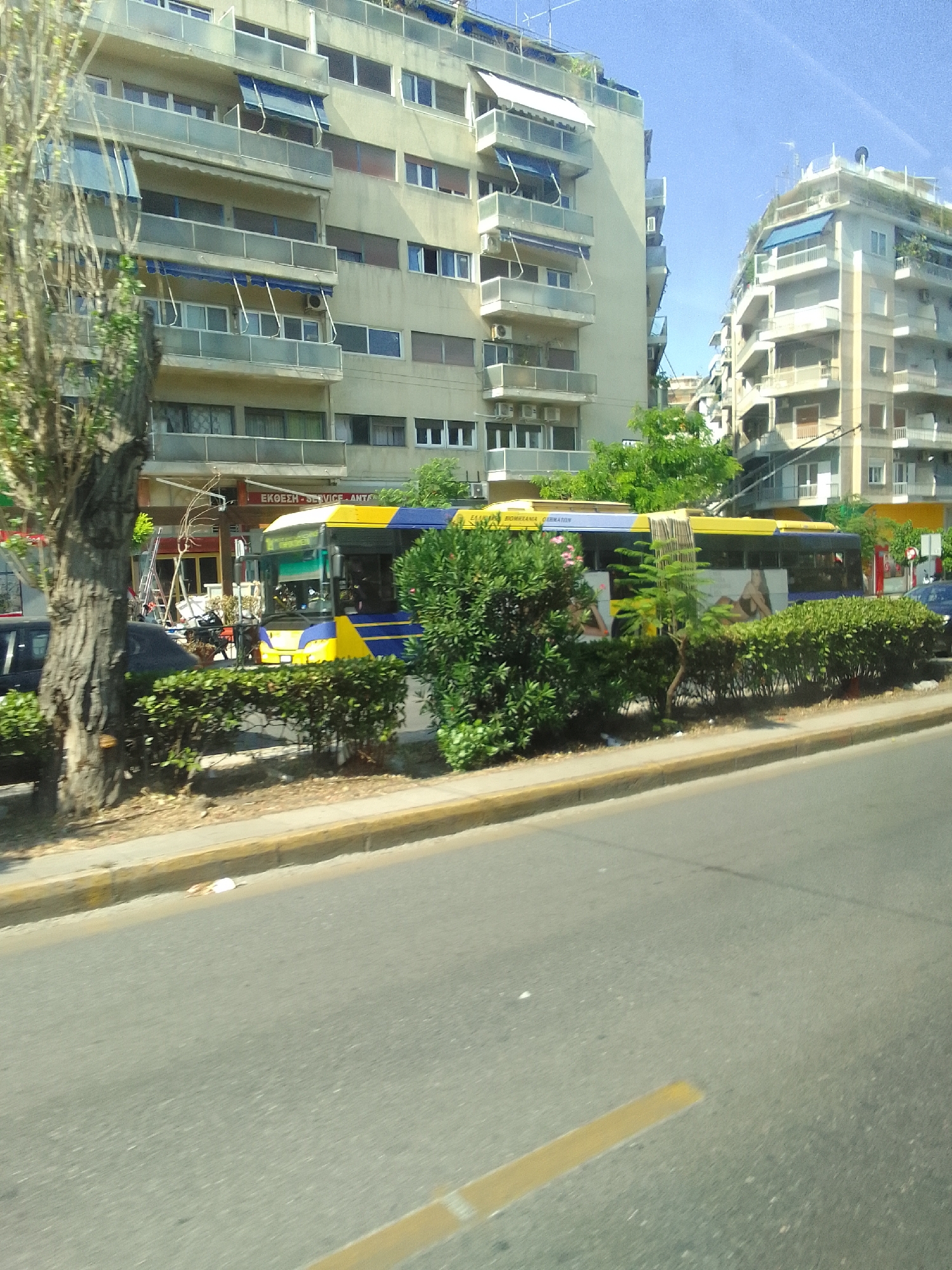 A bright yellow and blue bus is parked on a street in Athens, Greece. The bus has an advertisement on its side featuring a woman. The bus is positioned in front of a row of buildings that appear to be apartment complexes, with balconies and awnings. The buildings are light-colored, and the sky is a clear blue, suggesting a sunny day. To the left of the bus, there is a large tree with dark bark and green foliage. A hedge runs along the sidewalk in front of the bus and buildings. There are also some red and yellow signs visible on the building behind the bus. The street in the foreground is paved, with a yellow line marking the edge of the road. The scene appears to be a typical urban environment during daylight. There is some text visible on the bus, but it is difficult to read clearly.