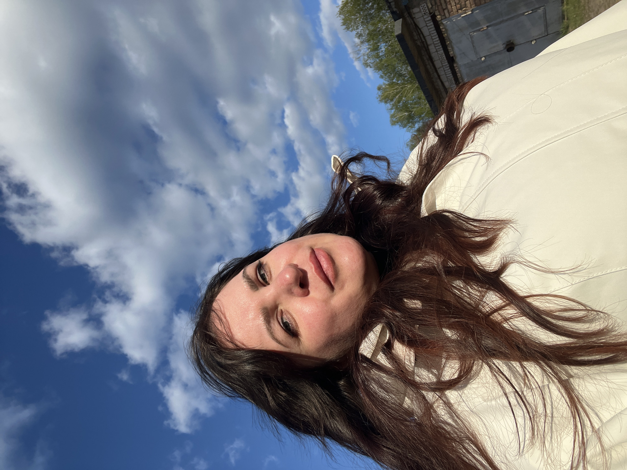 The image is a low-angle, slightly tilted selfie of a woman with long, dark hair wearing a white jacket. Her face is visible, showing facial features and skin texture. The background features a bright blue sky with fluffy white clouds. A corner of a brick building and some green trees are visible in the upper right corner. The scene appears to be outdoors on a sunny day. The geographical location is not discernible from the image. There is no definitive information in the picture that identifies the city as Noviy, Russia.
