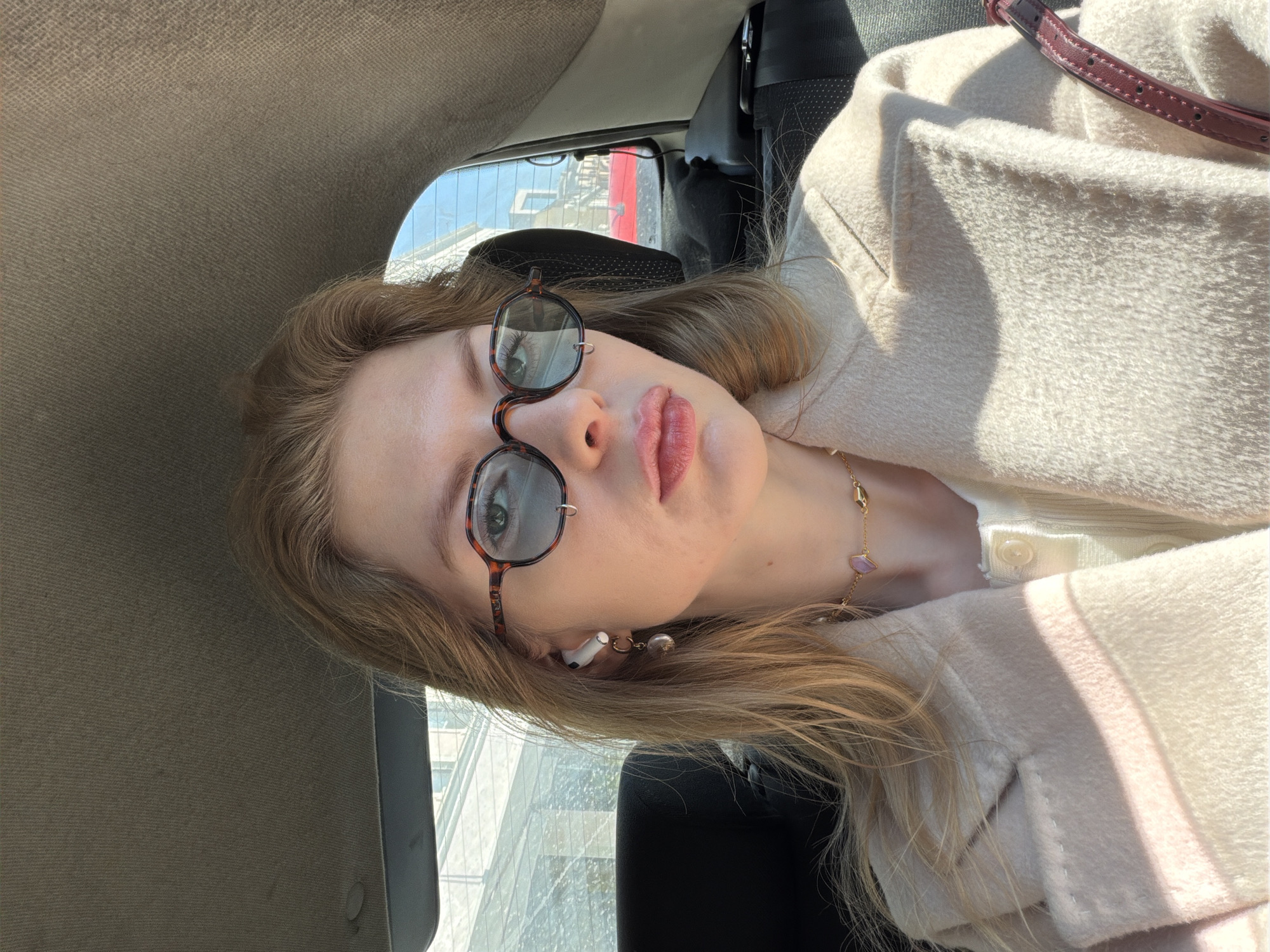 The image is a selfie taken by a young Caucasian woman with blonde hair while inside a vehicle, presumably a car. She is wearing round tortoise-shell sunglasses, a light-colored coat, and a delicate gold necklace with a small pendant. A white wireless earbud is visible in her left ear. The background suggests an urban setting, with a modern glass-fronted building visible through the car window.
Based on the architectural style of the building visible in the window, the location is likely within the city of Moscow, Russia. The building appears to have a contemporary design with reflective glass panels, a feature common in Moscow's modern business and commercial districts.