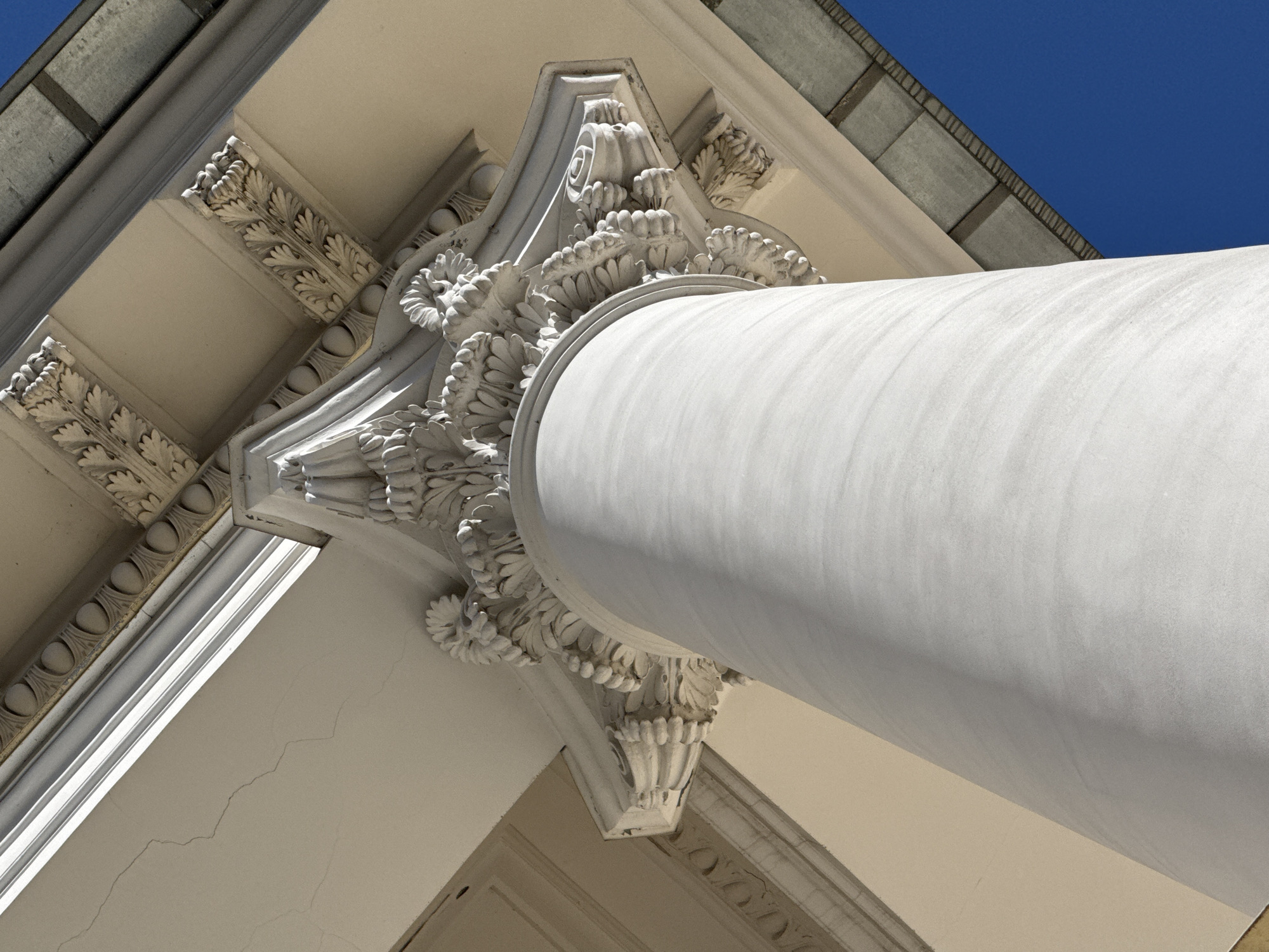 The image shows a low-angle view of a classical architectural detail, specifically the top of a column and its ornate capital, set against a clear blue sky. The column, which is cylindrical and smooth, dominates the right side of the frame. It is bright white and partially obscured by a light shadow that emphasizes its curvature.
The column's capital is highly decorative, with layers of floral and leaf-like carvings in a lighter shade of white or cream. This capital connects the column to the building's structure above. Above the capital, there is a decorative entablature with alternating leaf and rosette patterns.
To the left, the building's wall is visible, painted in a similar light shade. There are architectural details, including a cornice with an egg-and-dart motif. The top left corner of the frame shows a section of the roof or upper structure, with gray, rectangular tiles.
The scene suggests a formal, monumental building of significant architectural merit. Considering the architectural style, the image could be located in Saint Petersburg, Russia, which is known for its classical and neoclassical architecture.