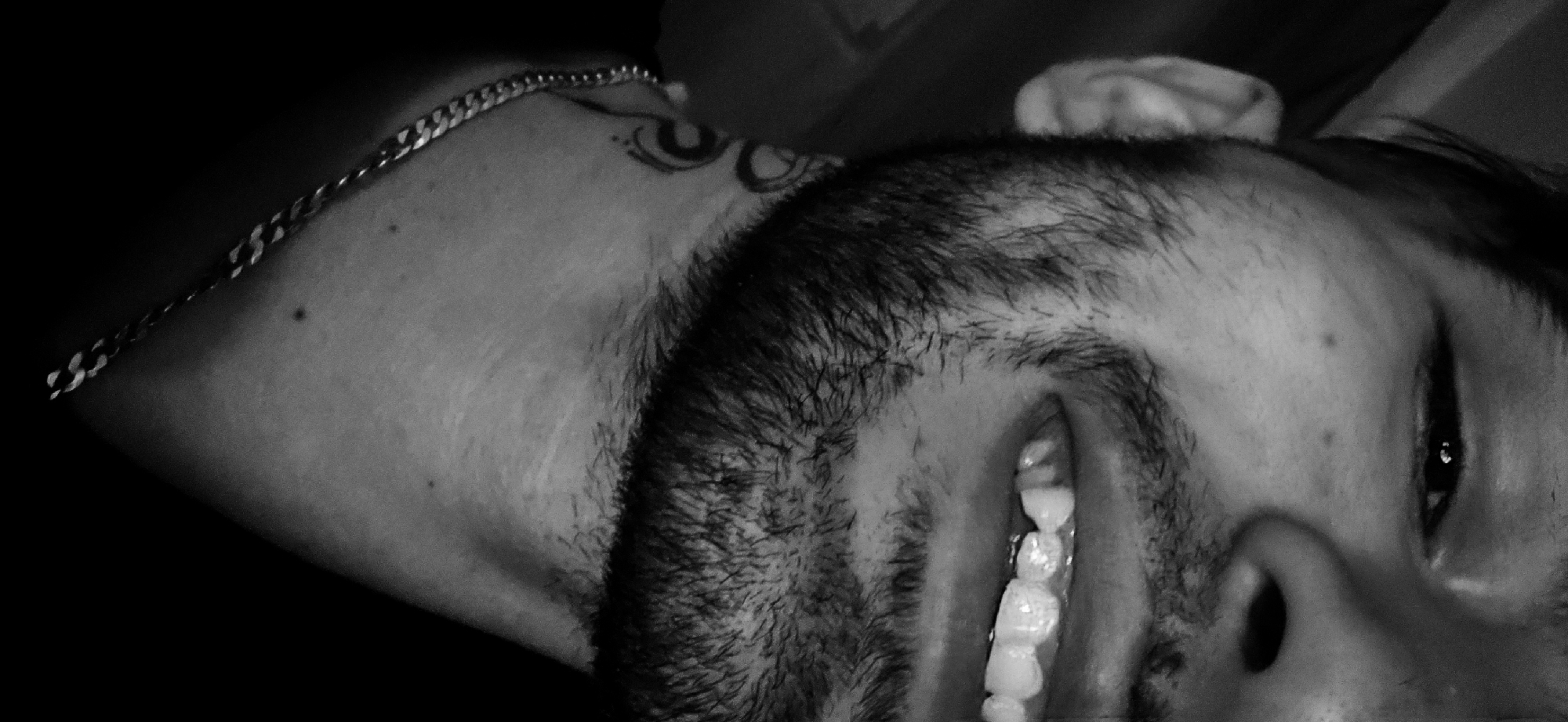 A black and white photograph depicts a man's face and neck, turned sideways. He is wearing a chain necklace. A tattoo is visible on his upper neck. His mouth is open, revealing his teeth. The background is dark and indistinct. The image provides no information about the city of Plovdiv, Bulgaria.
