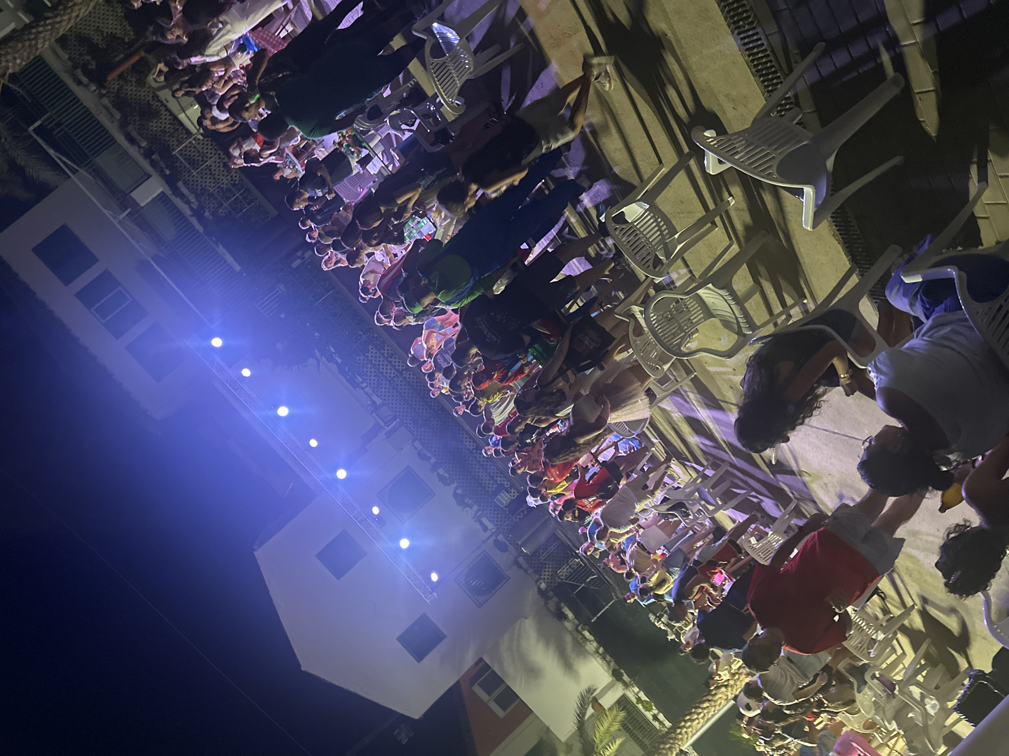 The image depicts an outdoor event at night, likely in Blanca, Spain, given the context. A crowd of people, varying in age and attire, are gathered, with many standing and some seated in white plastic chairs. The scene is illuminated by bright blue stage lights, casting long shadows and creating a vibrant, energetic atmosphere. In the background, a multi-story building with windows and balconies is visible, suggesting an urban or resort setting. Some individuals appear to be dancing or socializing, while others are observing the event. The overall impression is that of a lively outdoor concert or festival.