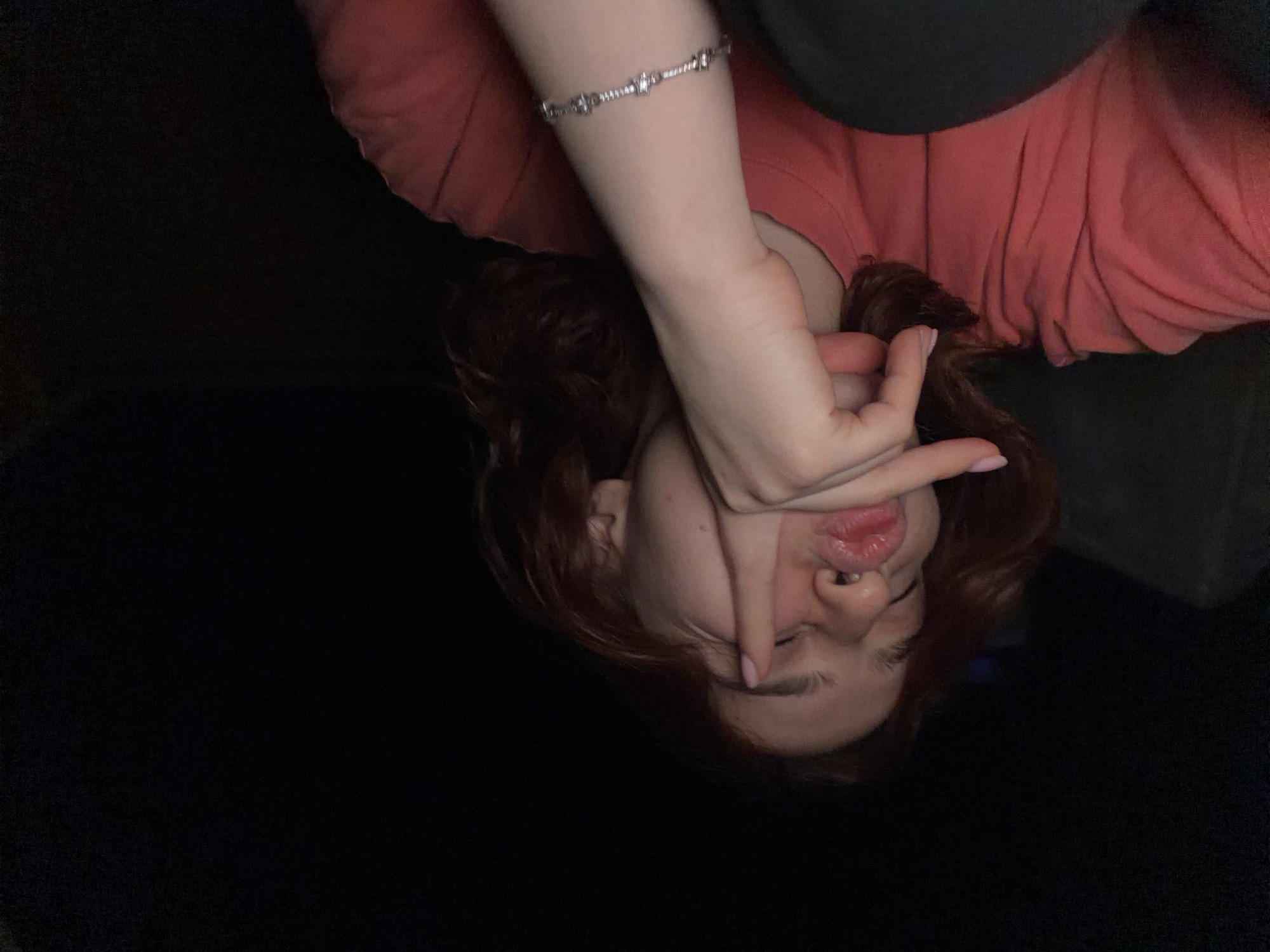 The image is a close-up, low-light selfie of a young person with reddish-brown hair. They are wearing a coral-colored t-shirt and a black garment. The person's eyes are closed, and they are puckering their lips, with their right hand raised to their face, touching their lips and cheek. A silver bracelet with small charms is visible on their left wrist. The background is dark and indistinct, suggesting an indoor or nighttime setting. There are no discernible elements indicating a specific location within Strasbourg, France.