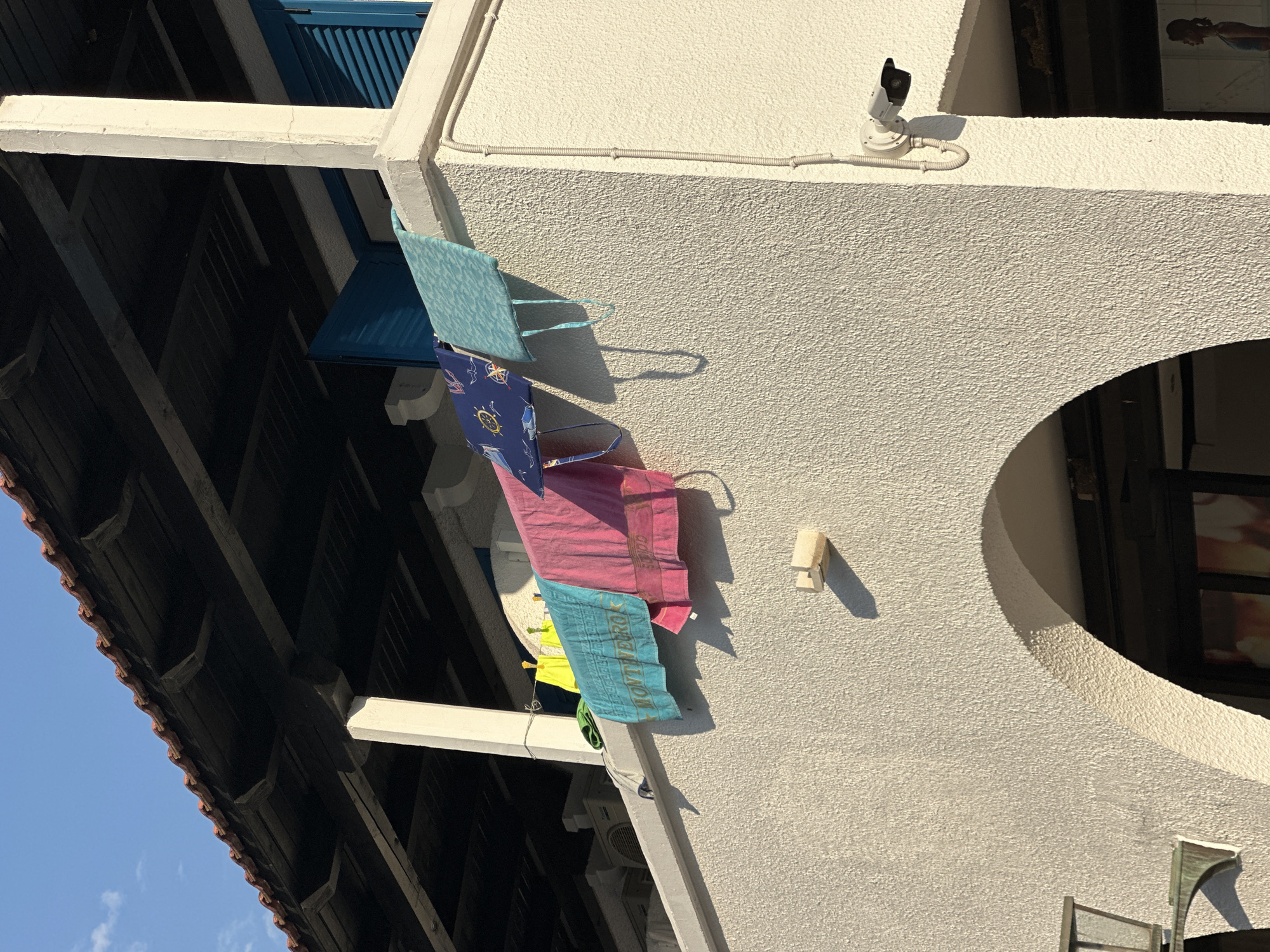 The image shows a white building with a tiled roof and a covered balcony. Several towels and bags are hanging on a clothesline on the balcony railing. The towels are in various colors including blue, pink, and yellow. One of the blue towels has the word "MONTENEGRO" printed on it. In the background, a blue shuttered window is visible. A security camera is mounted on the exterior wall of the building. The scene appears to be in Budva, Montenegro, given the text on the towel.