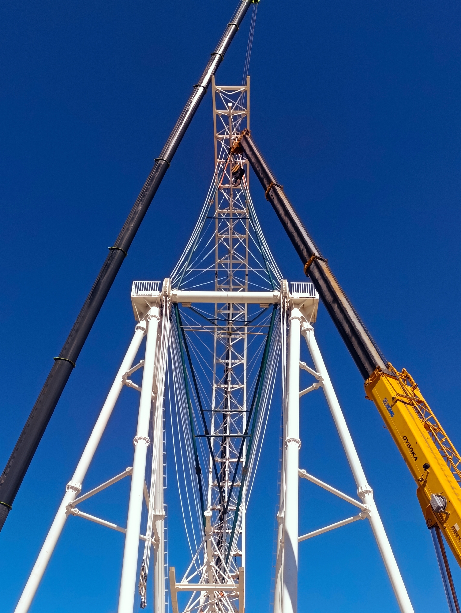 The image shows a low-angle, upward view of a large white steel structure, likely under construction, against a clear blue sky. Two large cranes are positioned on either side of the structure, their booms extending upwards. The crane on the left is black and appears to be a mobile crane, with its boom angled steeply upwards. The crane on the right is bright yellow and also angled upwards, with "XCMG QY50KA" visible on its boom, indicating its manufacturer and model. The white structure is complex, with numerous intersecting beams and supports. There are also white cables and some green cables supporting elements of the structure. A small platform with white railings is visible on the structure, and at least one person can be seen on this platform, appearing to be working. The scene suggests a large-scale construction project, possibly for a Ferris wheel or a tower, taking place in an outdoor, open environment. While the specific location within Naryn City, Kyrgyzstan is not visually discernible from the image alone, the presence of heavy construction equipment and a significant structure implies an industrial or development zone within the city.