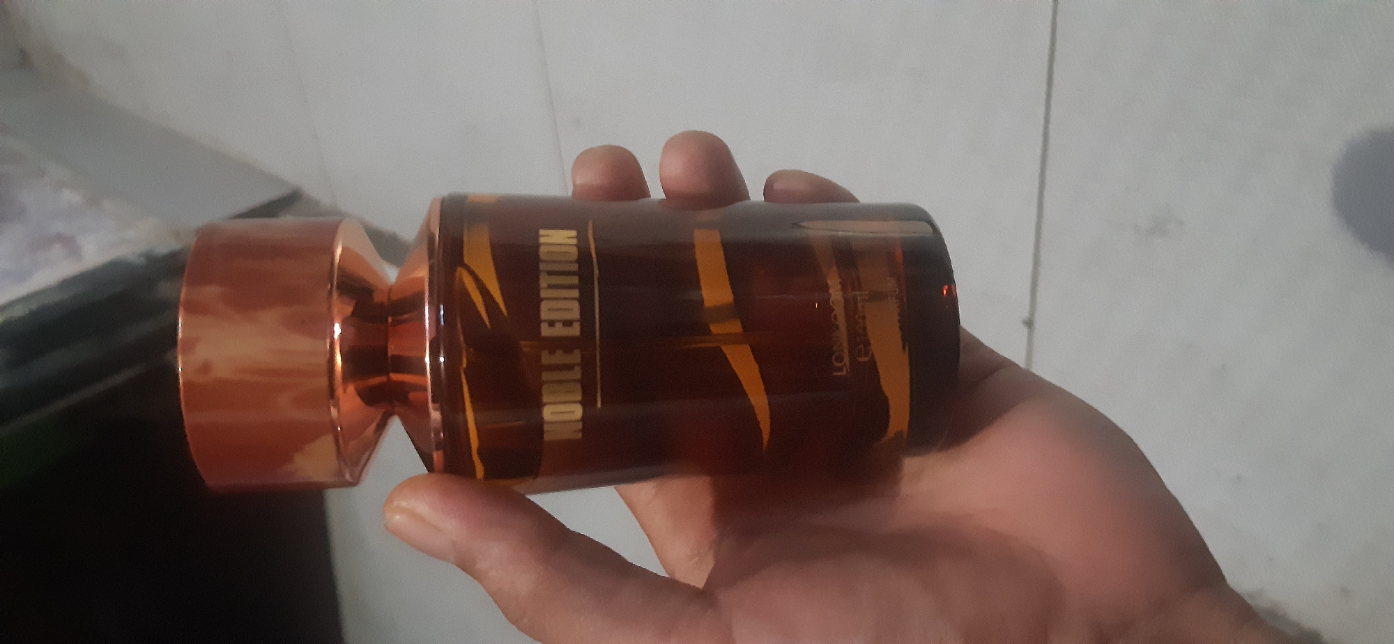 A hand holds a clear glass perfume bottle with amber-colored liquid. The bottle has a metallic copper-colored cap and a label with "NOBLE EDITION" printed in gold lettering. The background is a tiled floor. The location is not specified in the image or the provided crops.