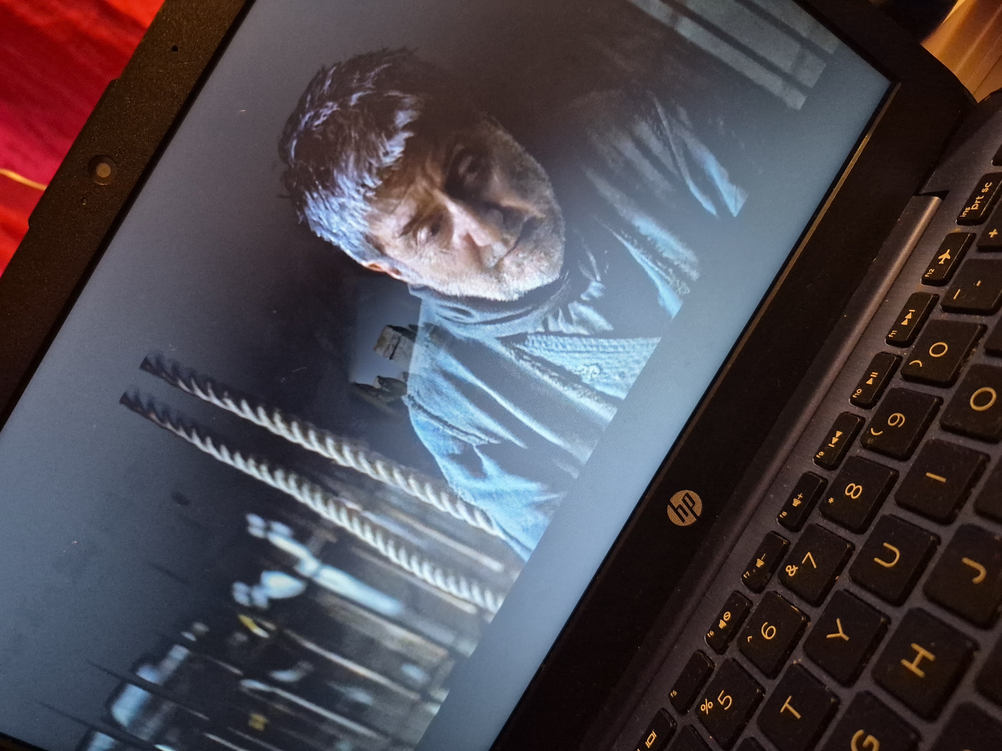 A close-up, high-angle shot shows the screen of a laptop displaying a scene from a movie or TV show. The screen shows a middle-aged man with grey hair and a beard, wearing a blue garment. He appears to be looking down with a somber expression. Behind him, ropes and wooden structures are visible, suggesting a ship or similar setting. The laptop keyboard is partially visible in the foreground, with various keys including letters, numbers, and function keys. The HP logo is visible on the laptop's casing. The image was taken indoors, likely in a domestic setting, and no specific location in Sandwell, United Kingdom, can be identified.