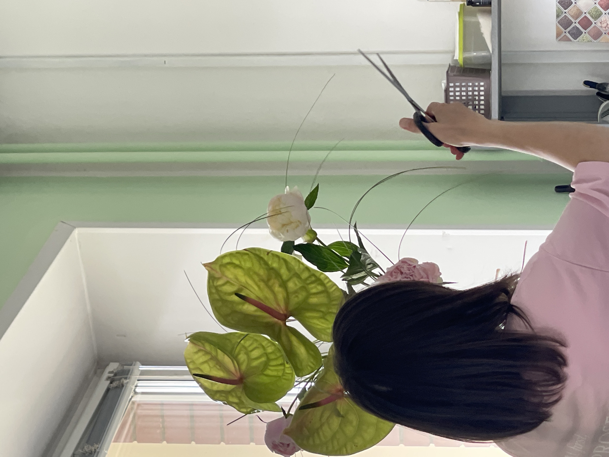 A person is arranging flowers. The person, with dark hair and wearing a pink shirt, is using scissors to trim a flower stem. The arrangement includes several bright green anthurium flowers and some delicate pink flowers, possibly peonies. The background appears to be an indoor setting with a window and pale green walls. The lighting suggests it is daytime.